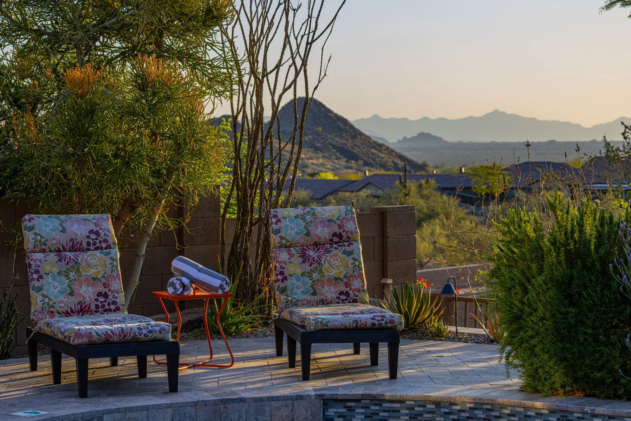 A backyard patio with two floral-patterned lounge chairs, rolled-up towels on a small orange table, and desert plants with mountains in the background during sunset.