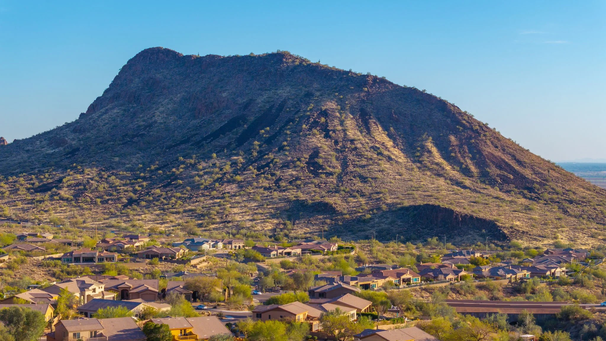 Residential neighborhood at the base of a mountain in a desert landscape with sparse vegetation and clear blue sky.