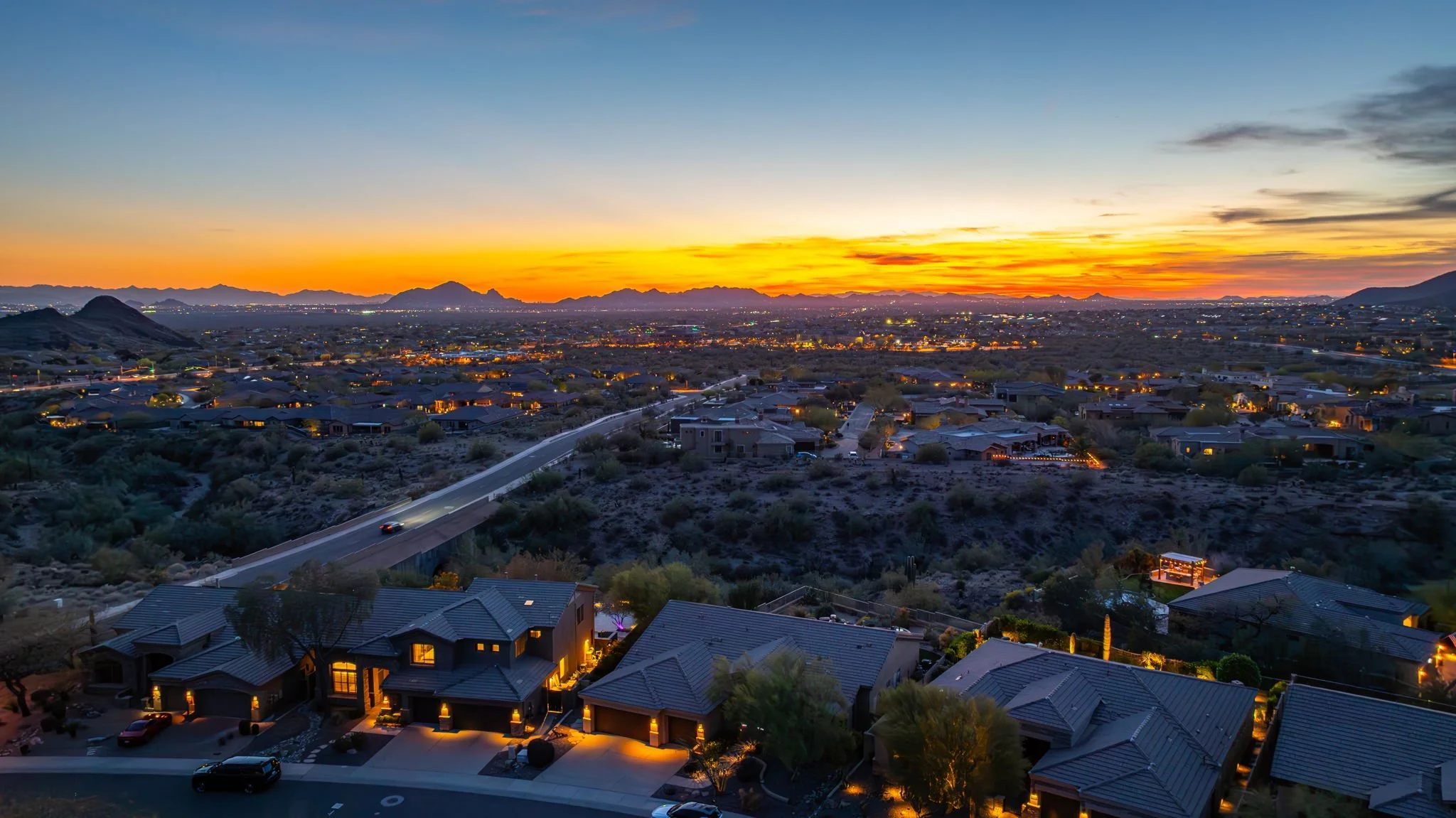 Aerial view of a suburban neighborhood at sunset with illuminated houses, streets, and a distant city skyline against a mountain range.