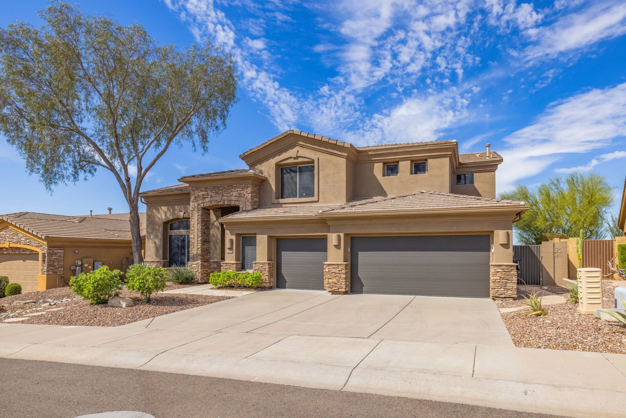 A two-story modern house with a desert landscape, large trees, and a three-car garage, under a blue sky with some clouds.
