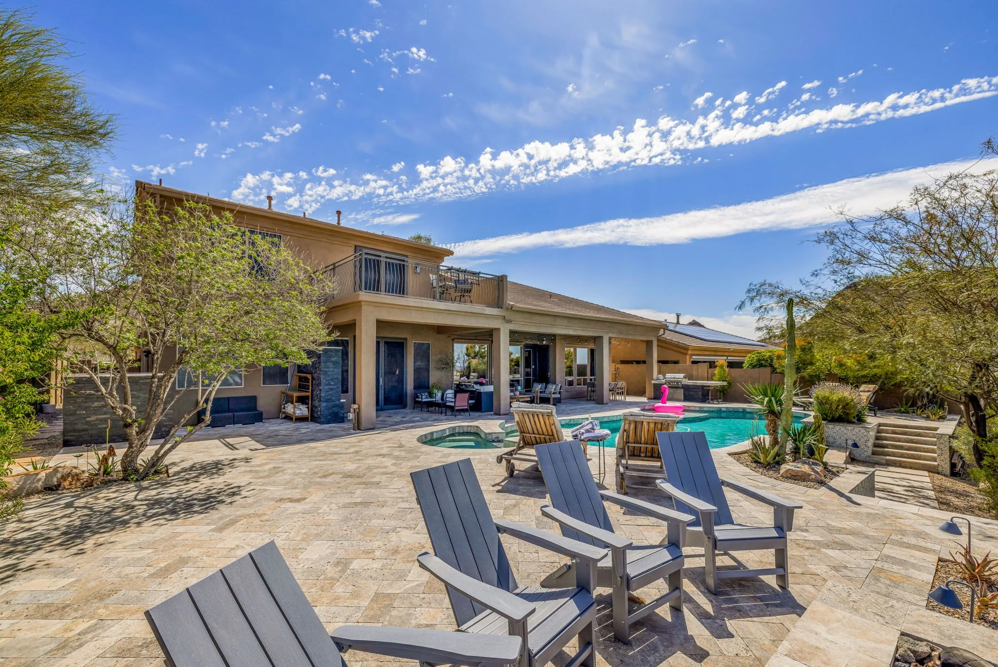 Backyard with a swimming pool, outdoor chairs, trees, and a two-story house with a patio under a blue sky.