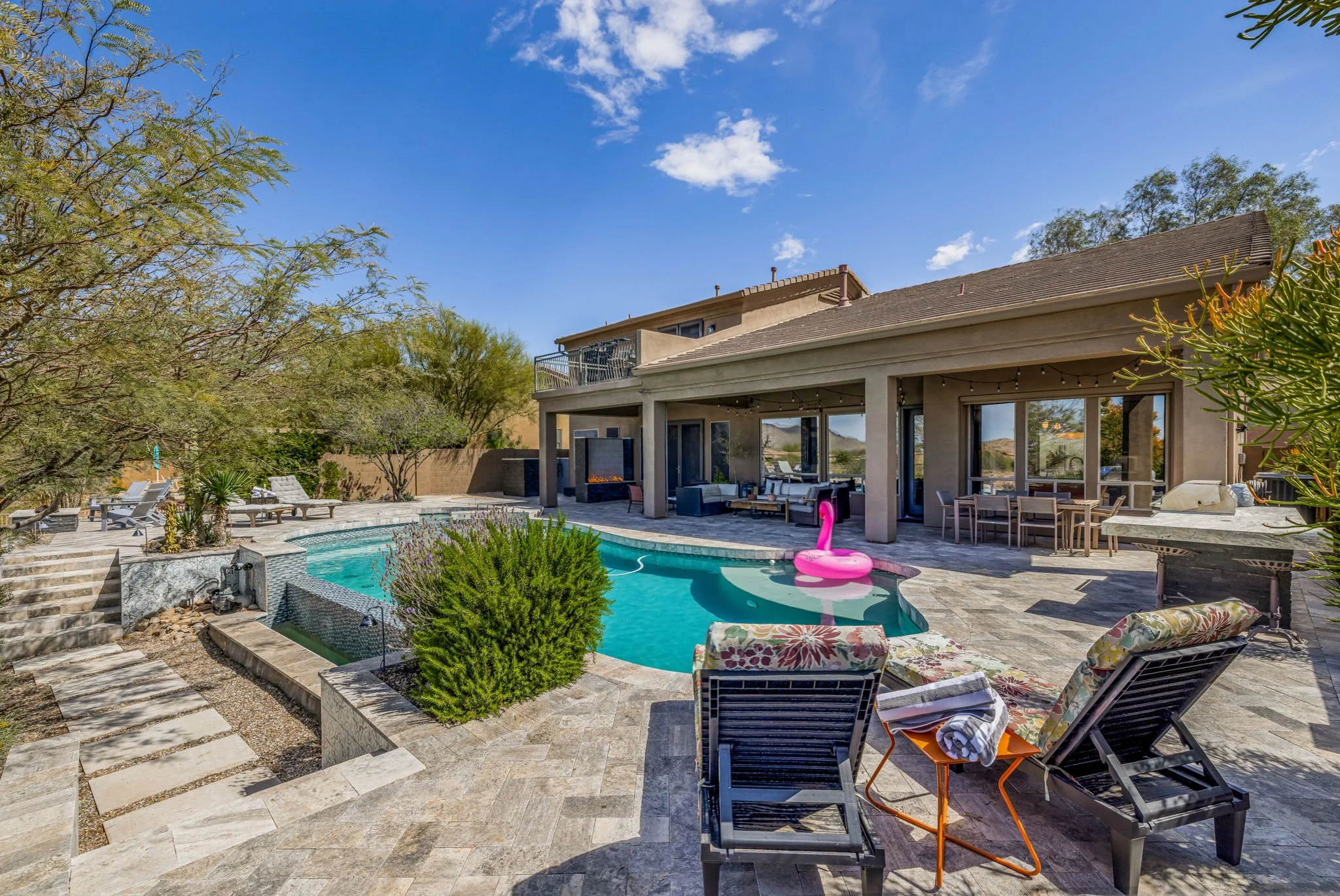 Residential backyard with a swimming pool, lounge chairs, patio furniture, and a pink flamingo float, surrounded by desert landscaping and a two-story house under a blue sky with scattered clouds.