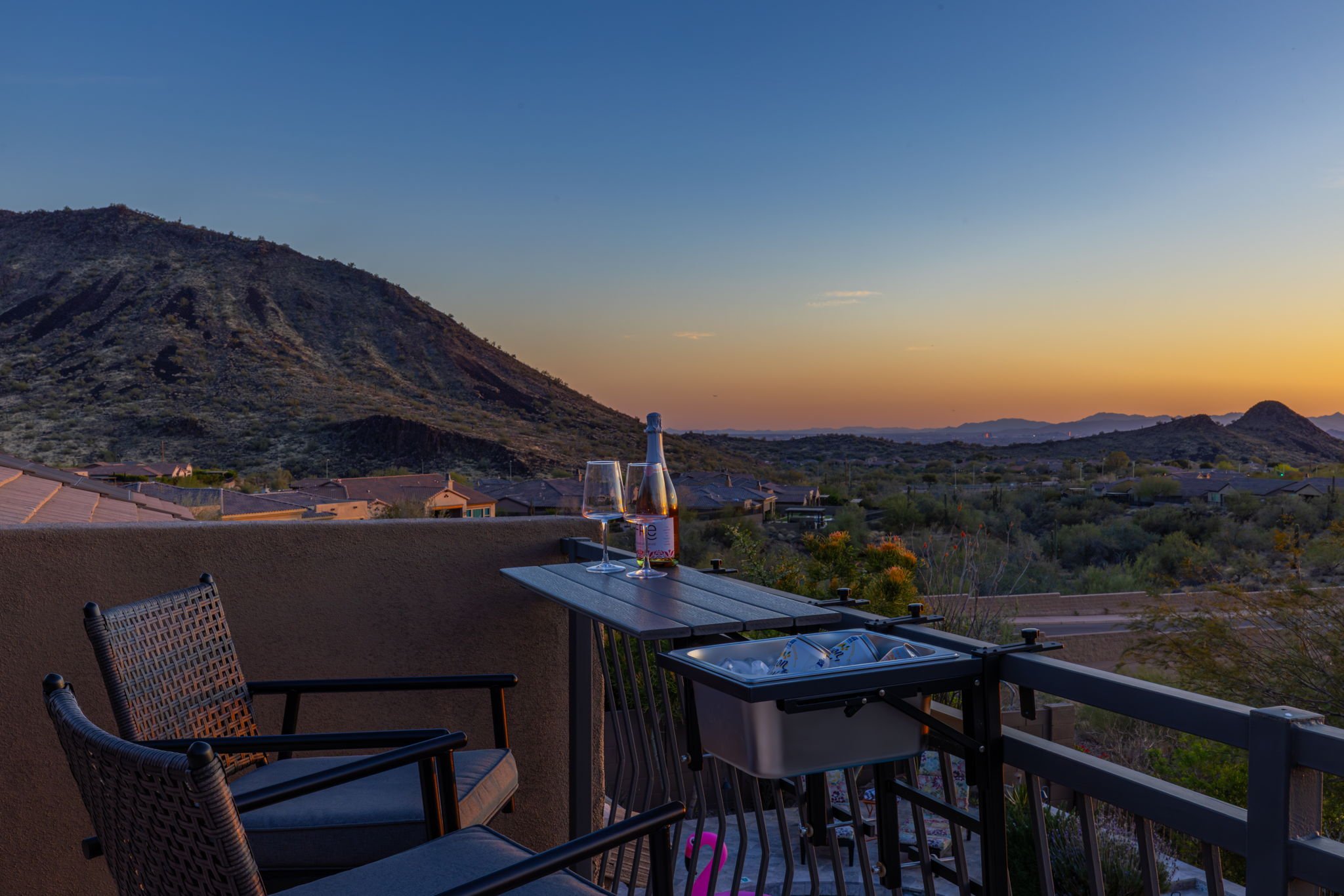 A balcony with two chairs, a small table with a bottle of wine, two glasses, and a cooler, overlooking a desert landscape with mountains and a sunset sky.