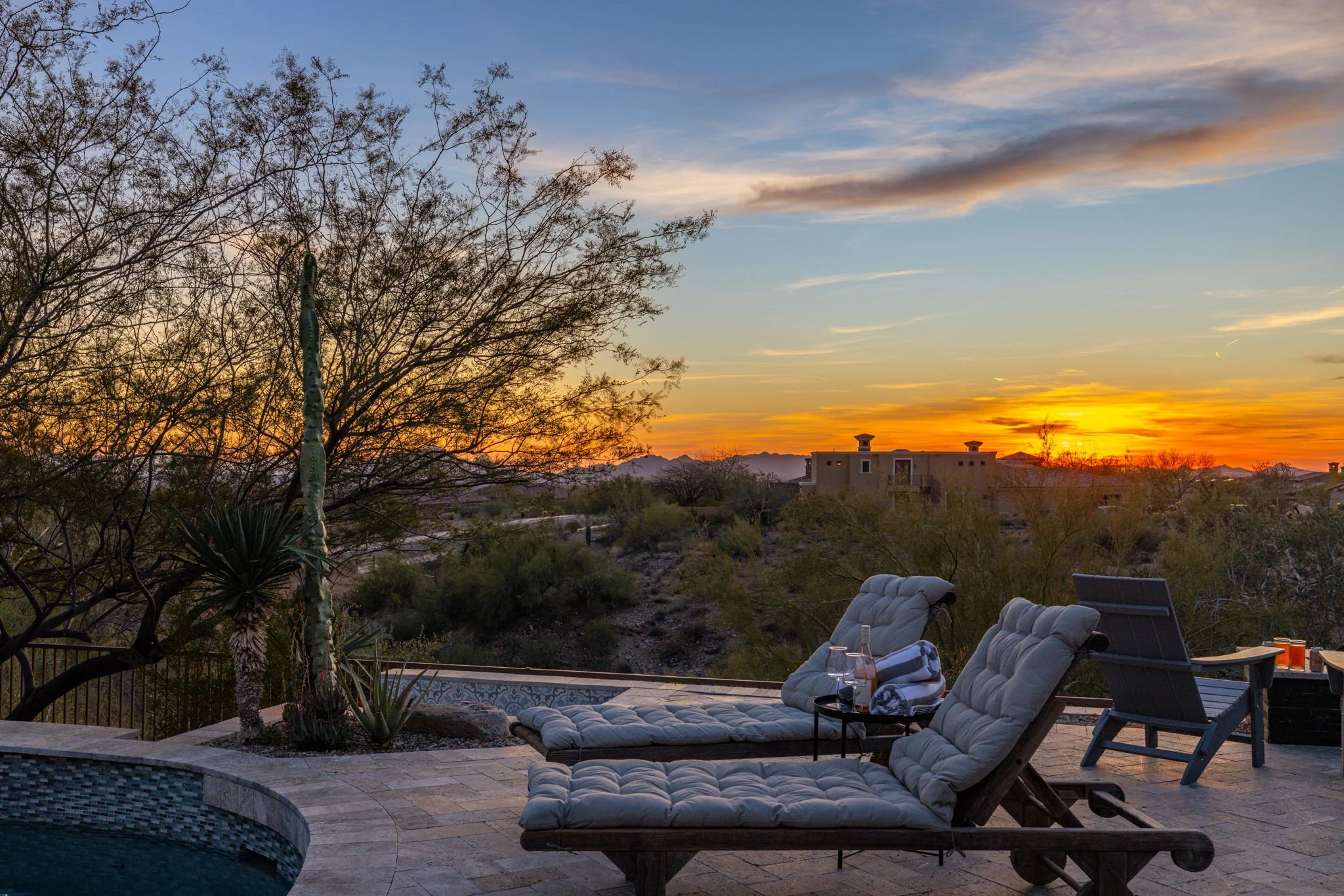 Patio in desert landscape during sunset with lounge chairs, table with drinks, and desert plants.