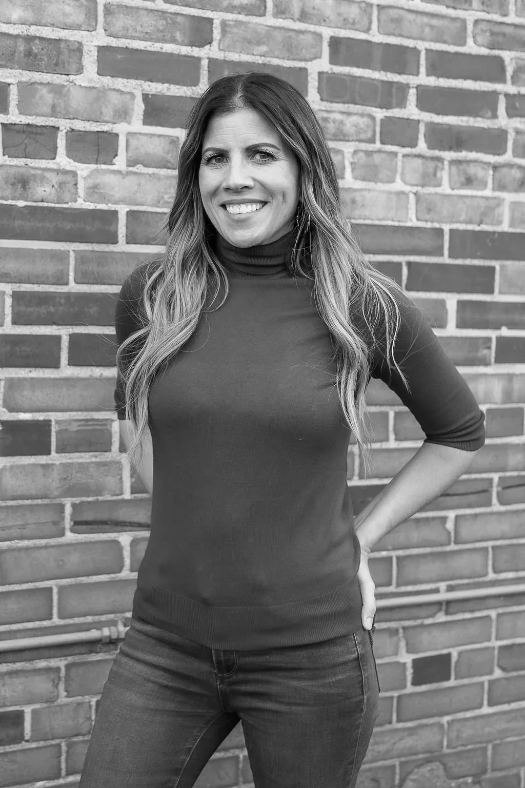 A woman with long wavy hair smiling and posing with one hand on her hip against a brick wall.