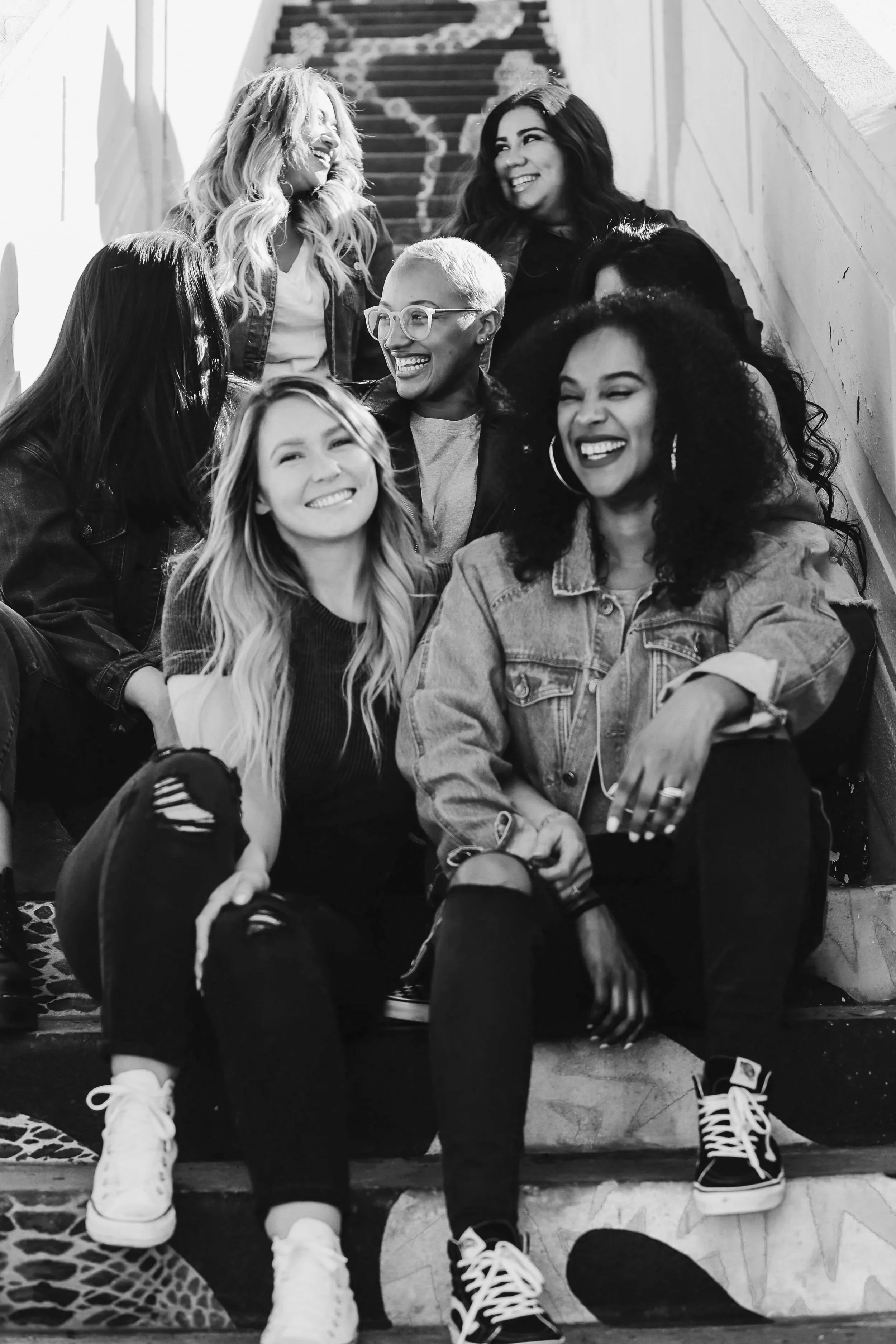 Group of six women sitting on stairs, smiling and laughing together, in a casual setting.