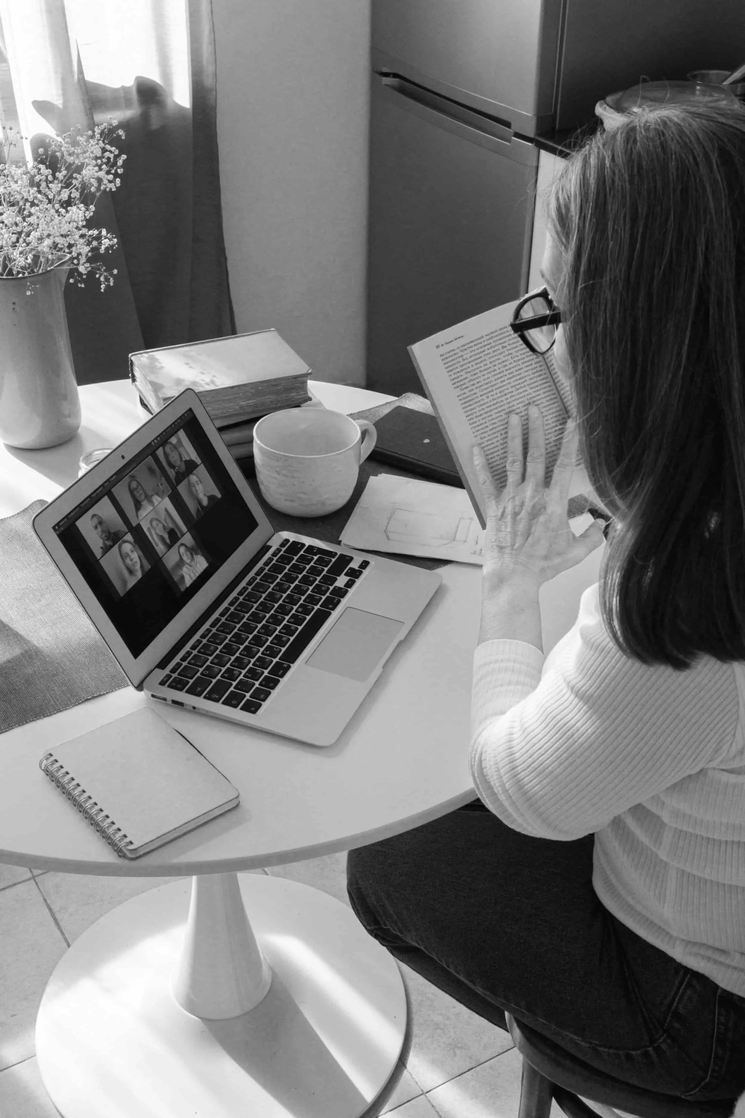 Woman on virtual coaching call at round table with laptop, book, mug, notebook, and flowers in natural light.
