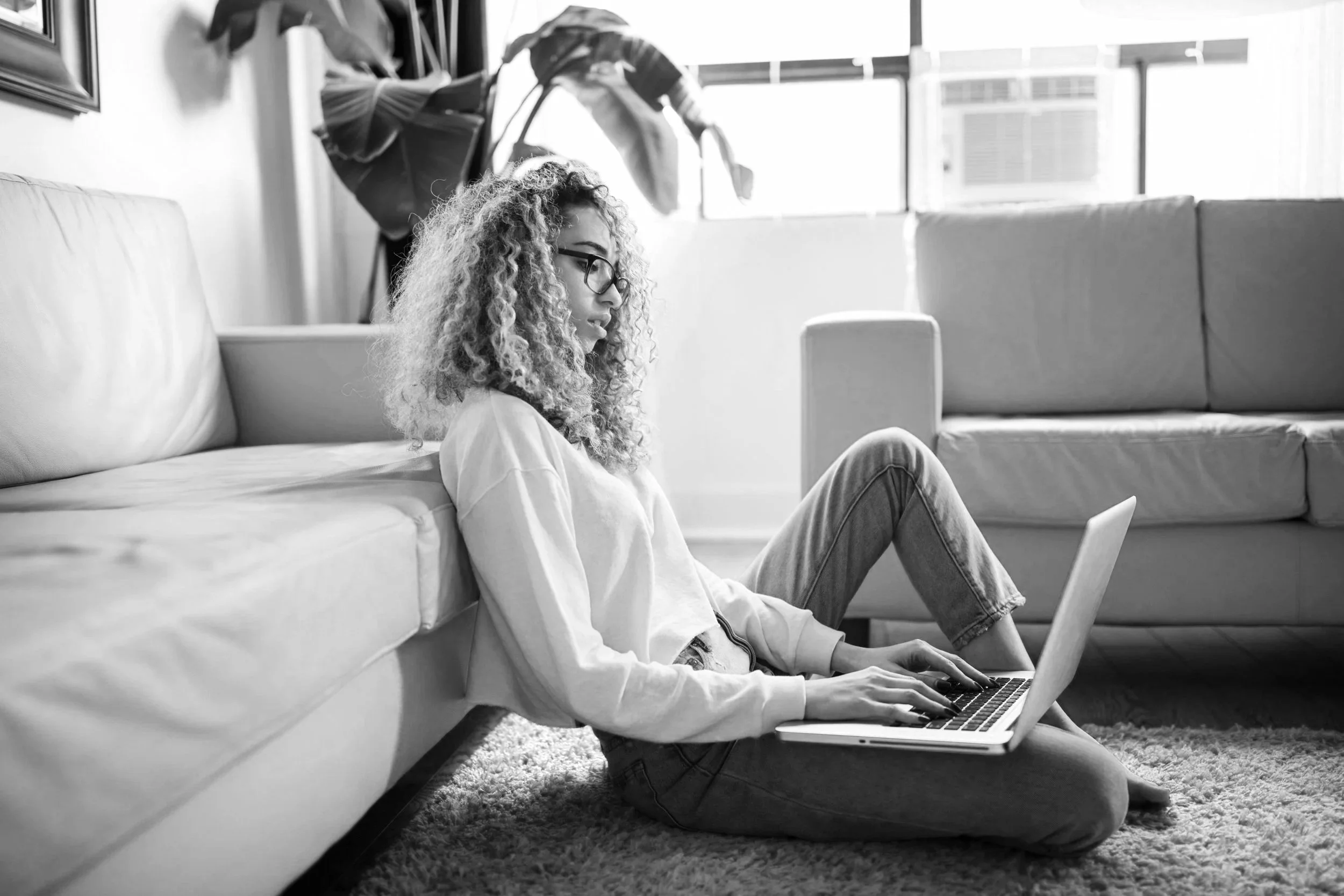 A young woman with curly hair and glasses sitting on the floor, leaning against a couch, working on her laptop with another device nearby in a brightly lit room.