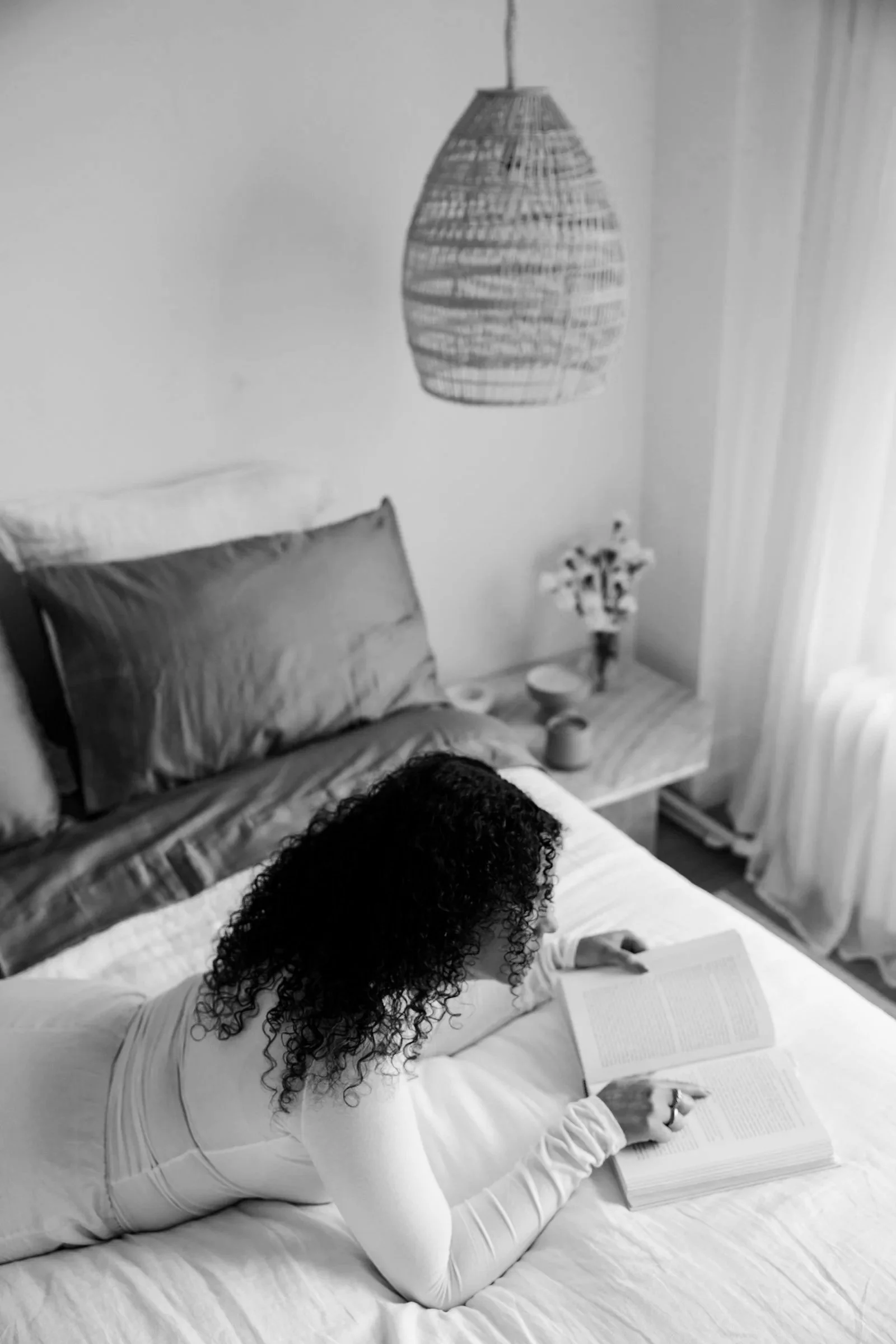 A woman with curly hair is lying on her stomach on a bed, reading a book. She is wearing a white outfit. The bed has a pillow, and in the background, a nightstand with a flower vase and a lamp hanging from the ceiling