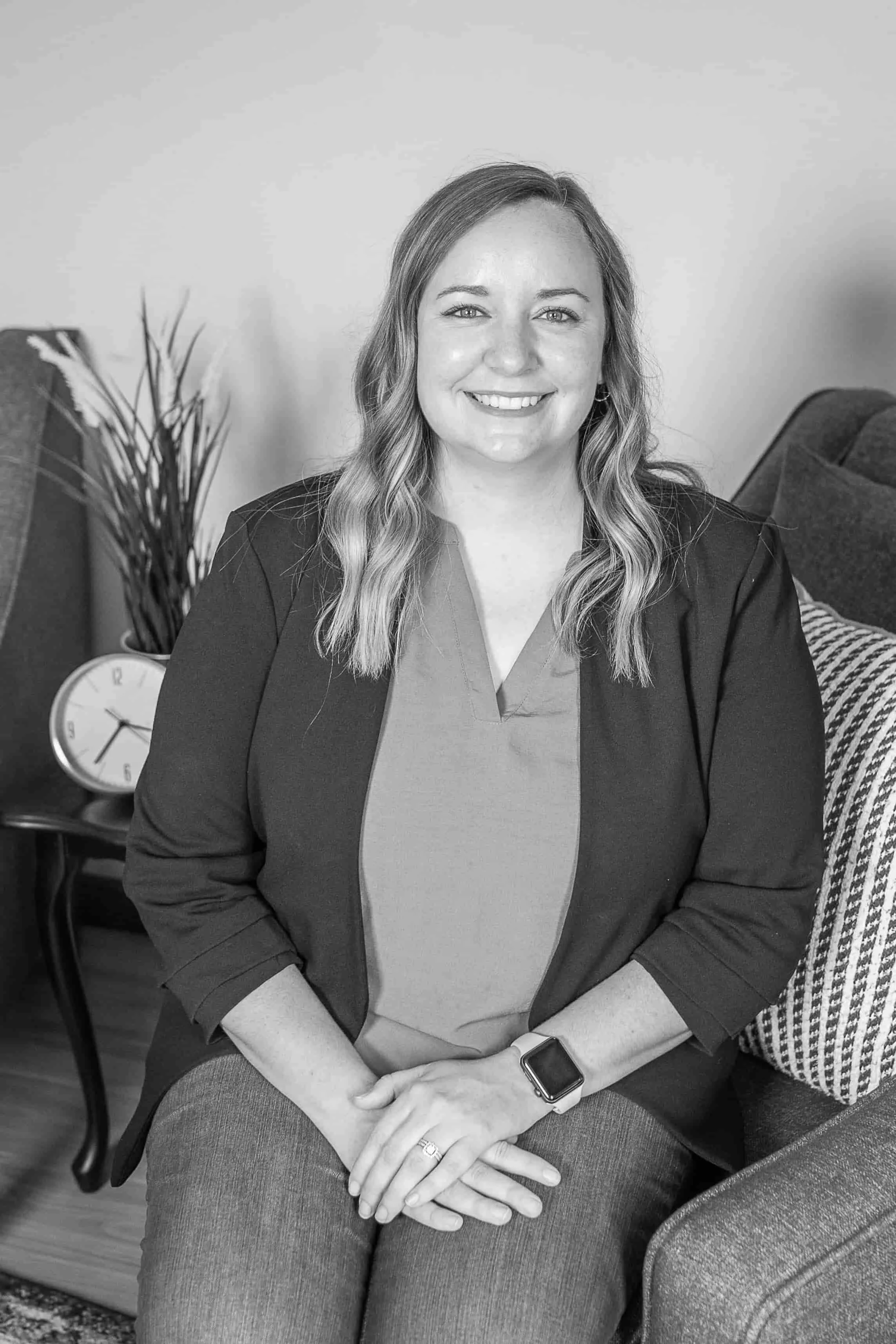 Ellie Blahnik, LMFT, smiles as she sits on a couch in a blazer and smart watch, with a clock and plant behind her.