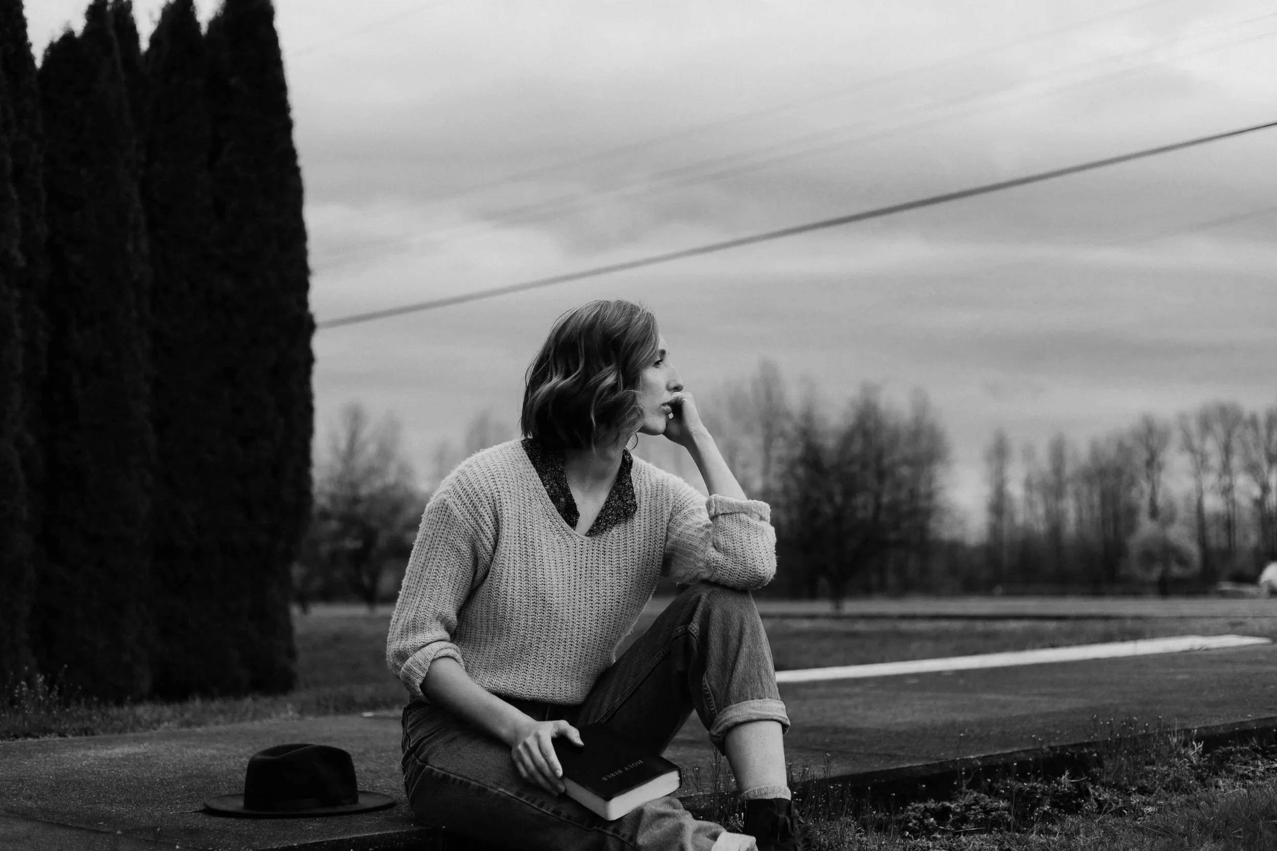 A black-and-white photo of a woman sitting on the grass near a large tree, with her hand on her face, looking thoughtfully into the distance. A hat and a book are placed on the ground beside her, with a landscape of trees and power lines in the background.