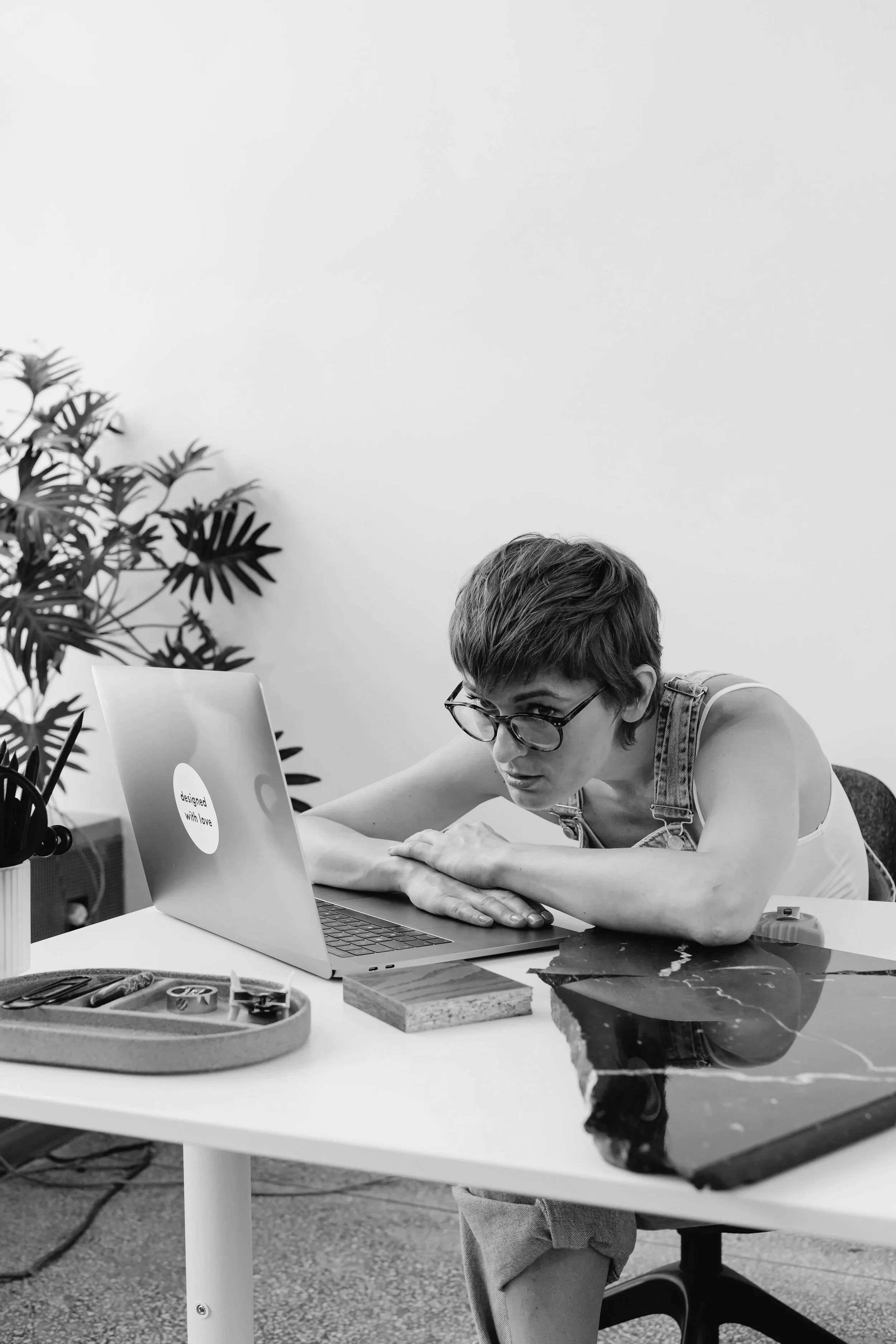A woman with short hair and glasses leaning over a table, working on a laptop in an office setting with plants, books, and a marble table.