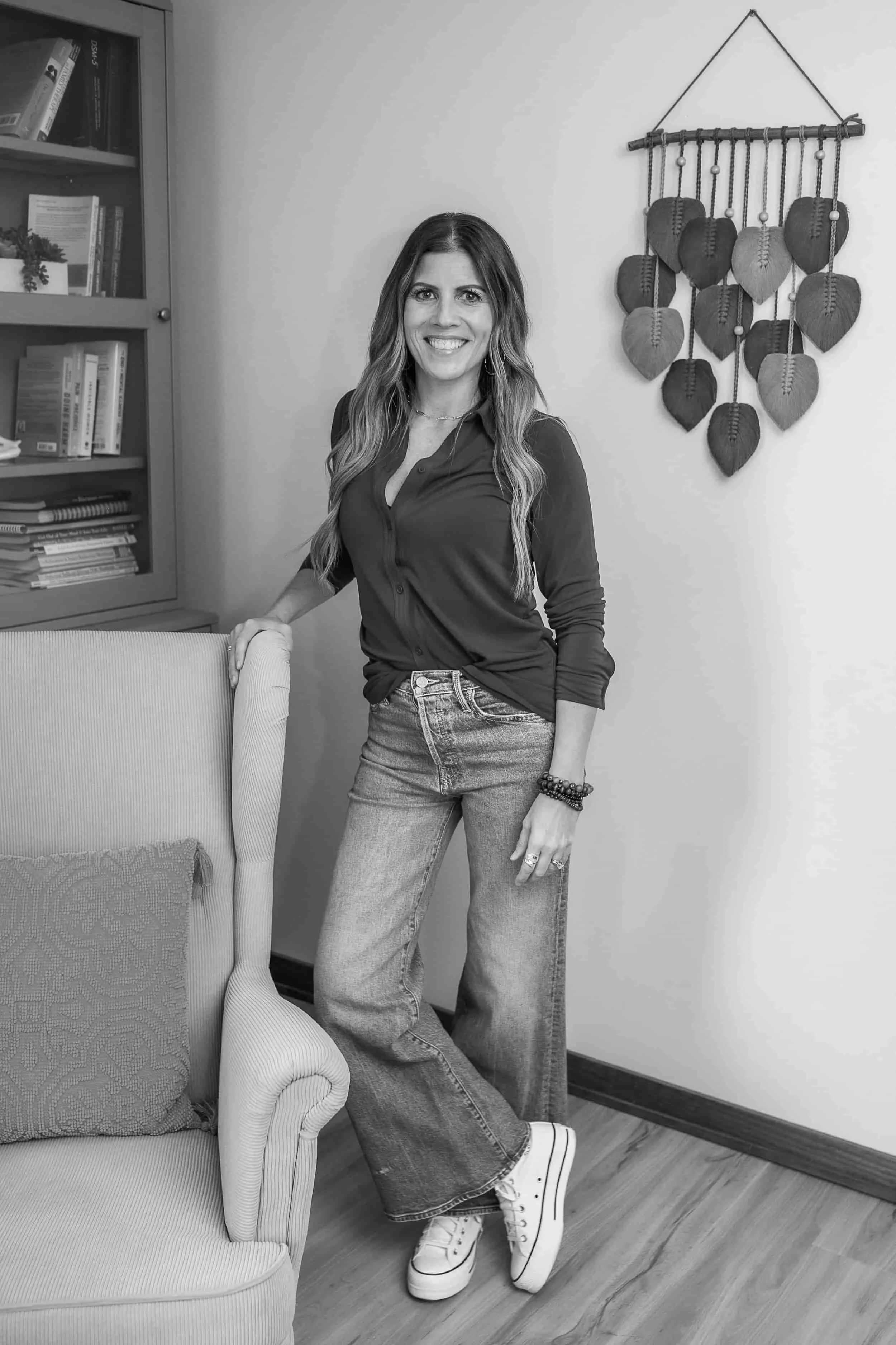 Angela Langer, LICSW, stands by an armchair, smiling in jeans and a dark top; wall art and bookshelf add charm. Black and white.
