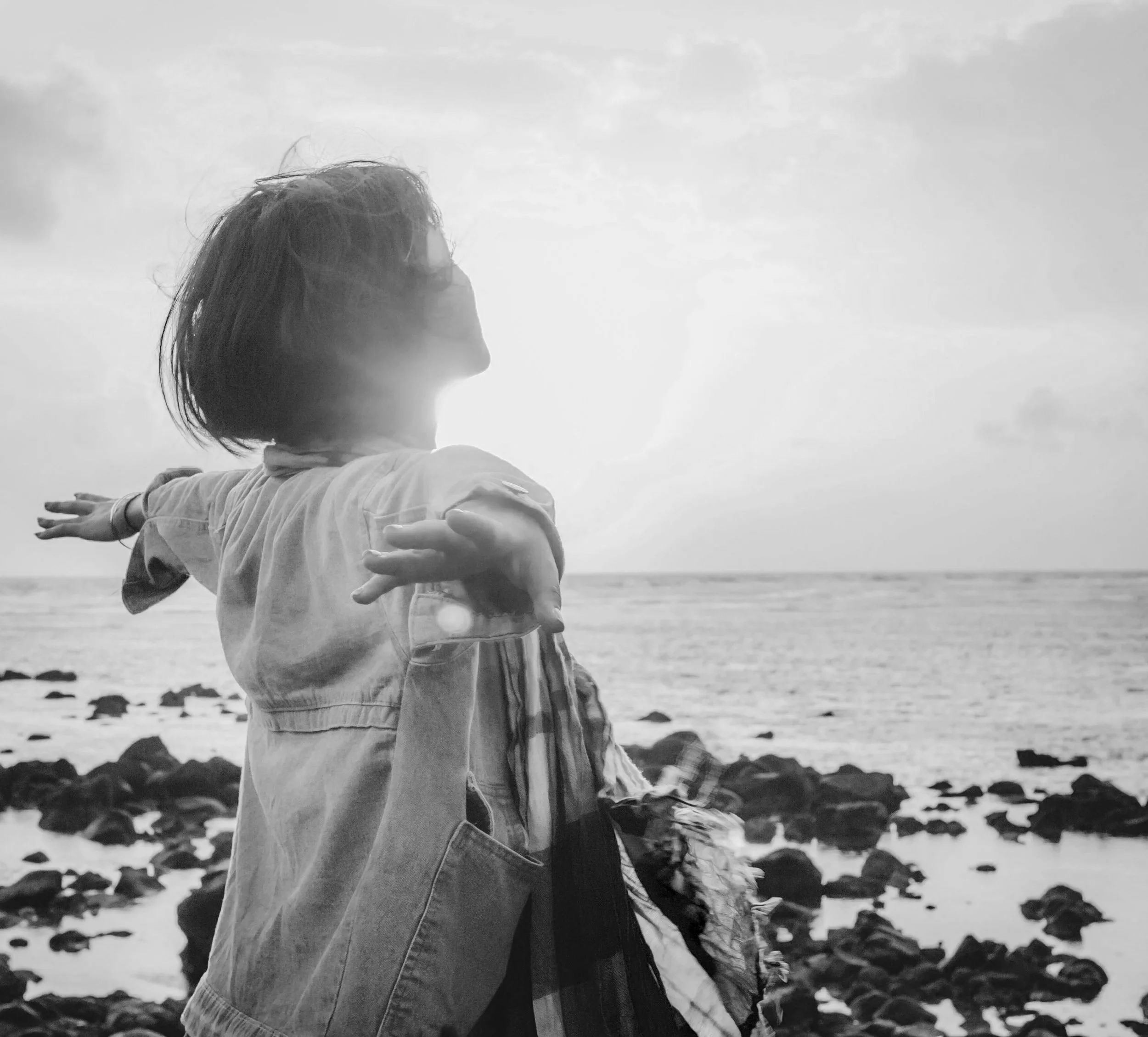 A black-and-white photo of a person standing outdoors near the water, with arms extended to the side, facing east with their profile visible. The person has medium-length hair and is wearing a long-sleeved shirt, with a natural, cloudy sky in the background.