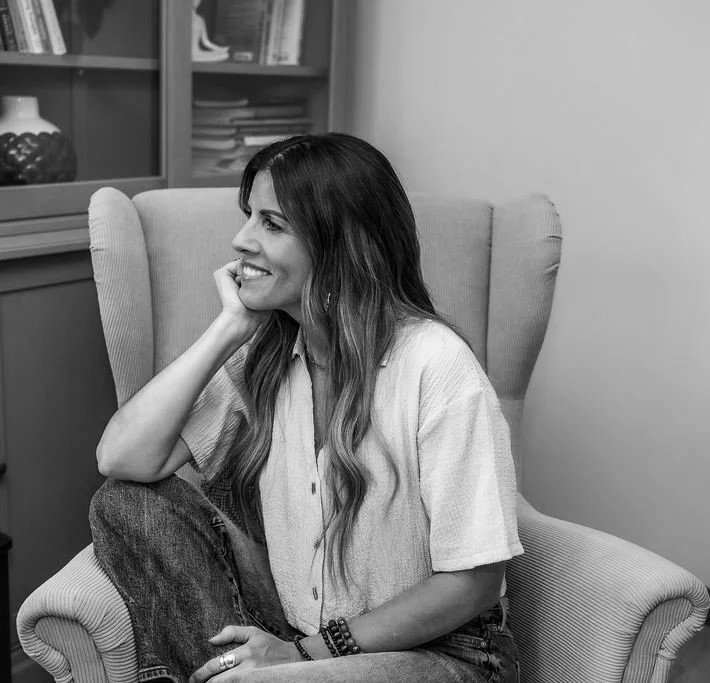 A woman with long, wavy hair smiling and sitting in a comfortable armchair, resting her chin on her hand, in a cozy indoor setting.