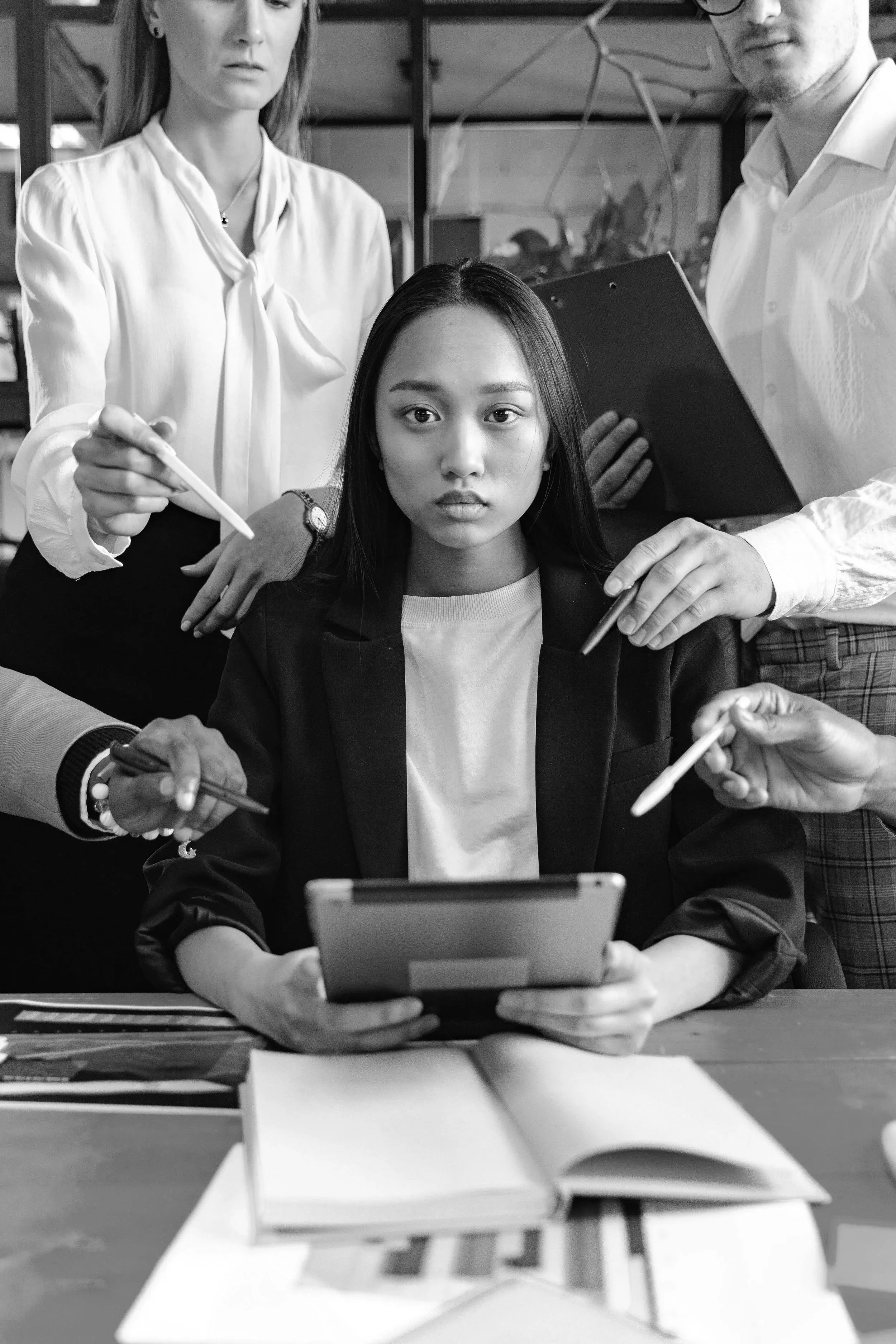 Black and white photo of a young woman sitting at a table, looking at a tablet, with hands holding styluses and about to work on the tablet. Behind her, three adults appear to be discussing or analyzing something, with two of them holding notebooks or papers. The setting looks like an office or a creative workspace.