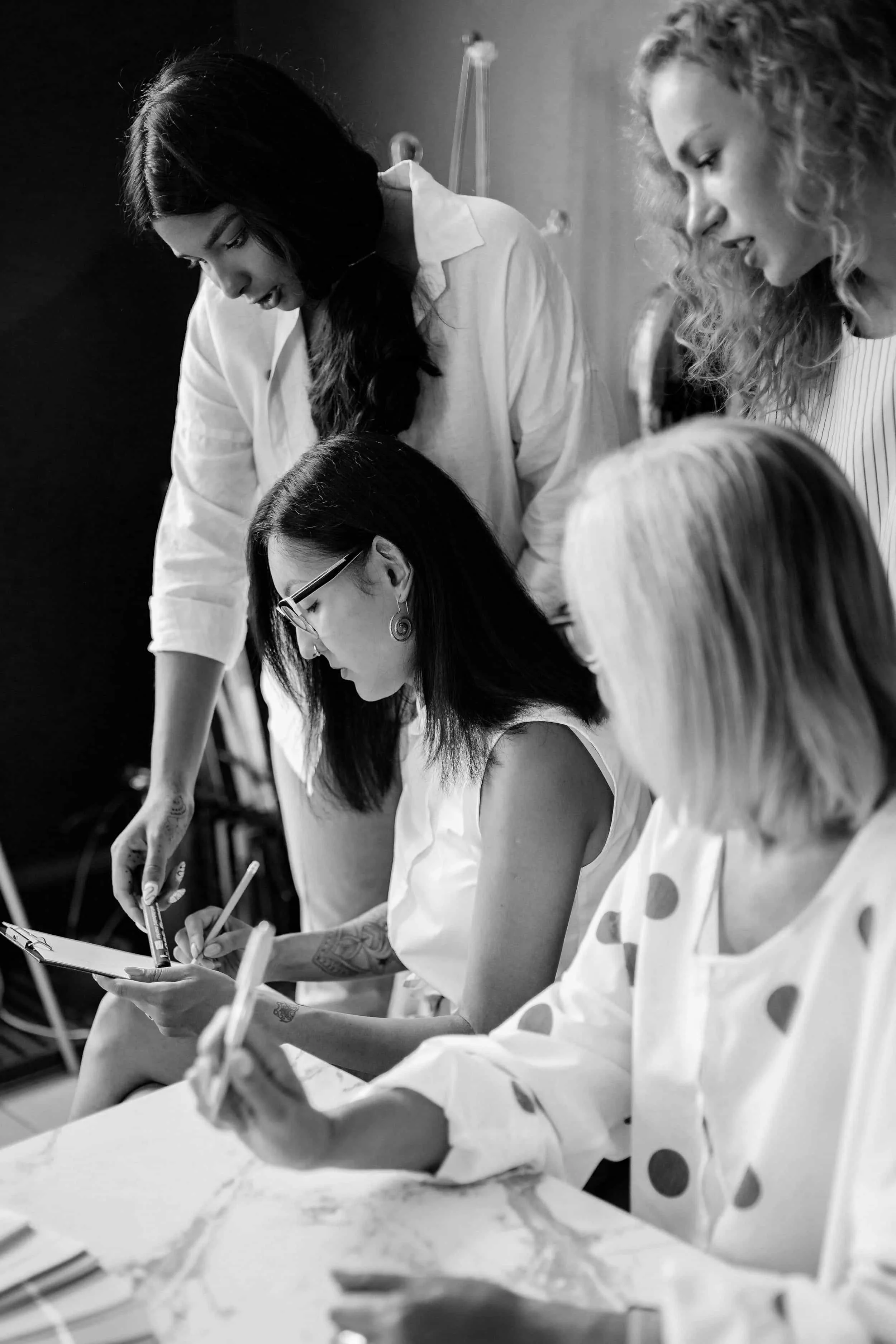 Four women collaborate at a table, discussing and writing notes, in a black and white photo.