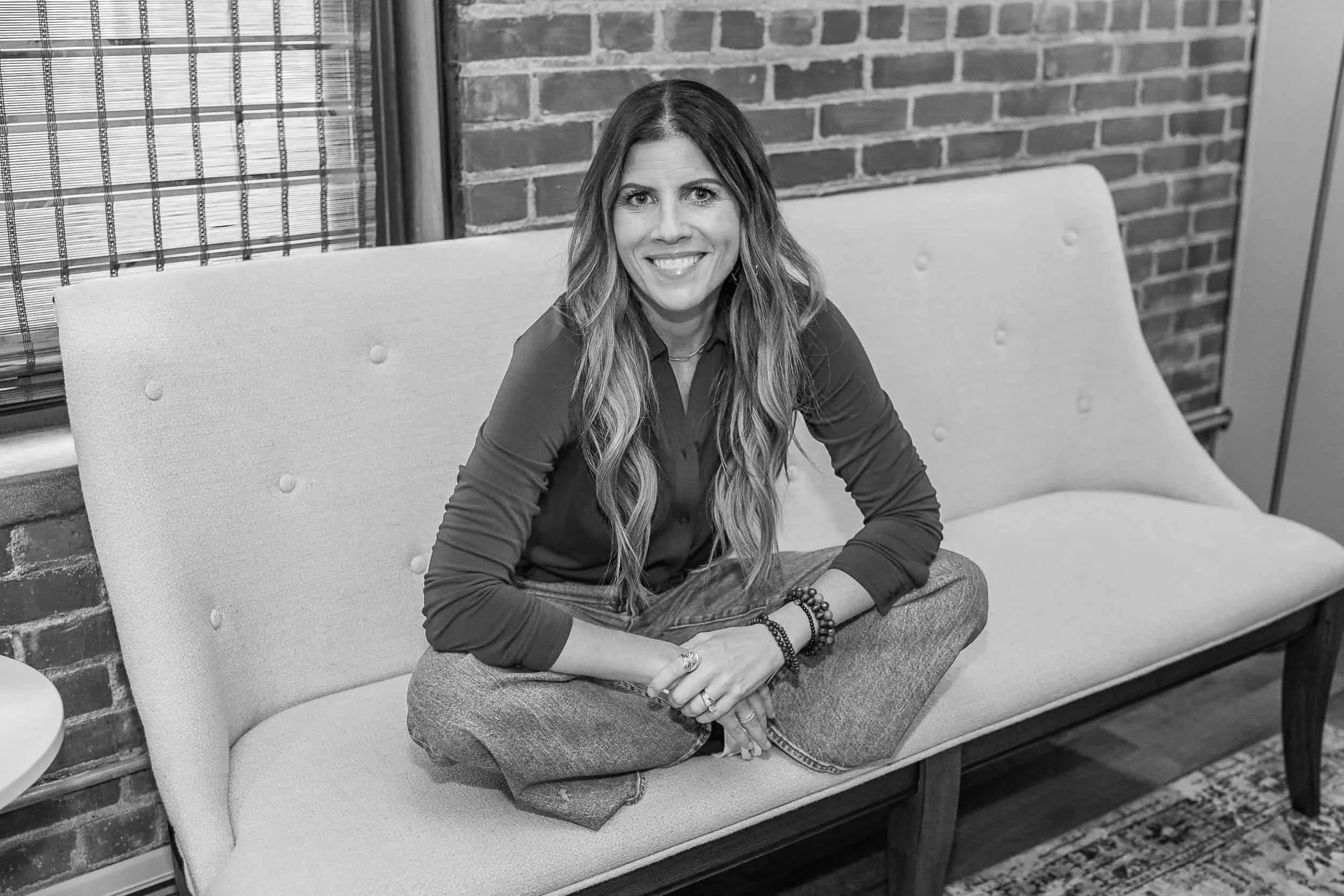 Angela Langer LICSW sits cross-legged on a cushioned bench, smiling in a brick-walled room. Black and white photo.