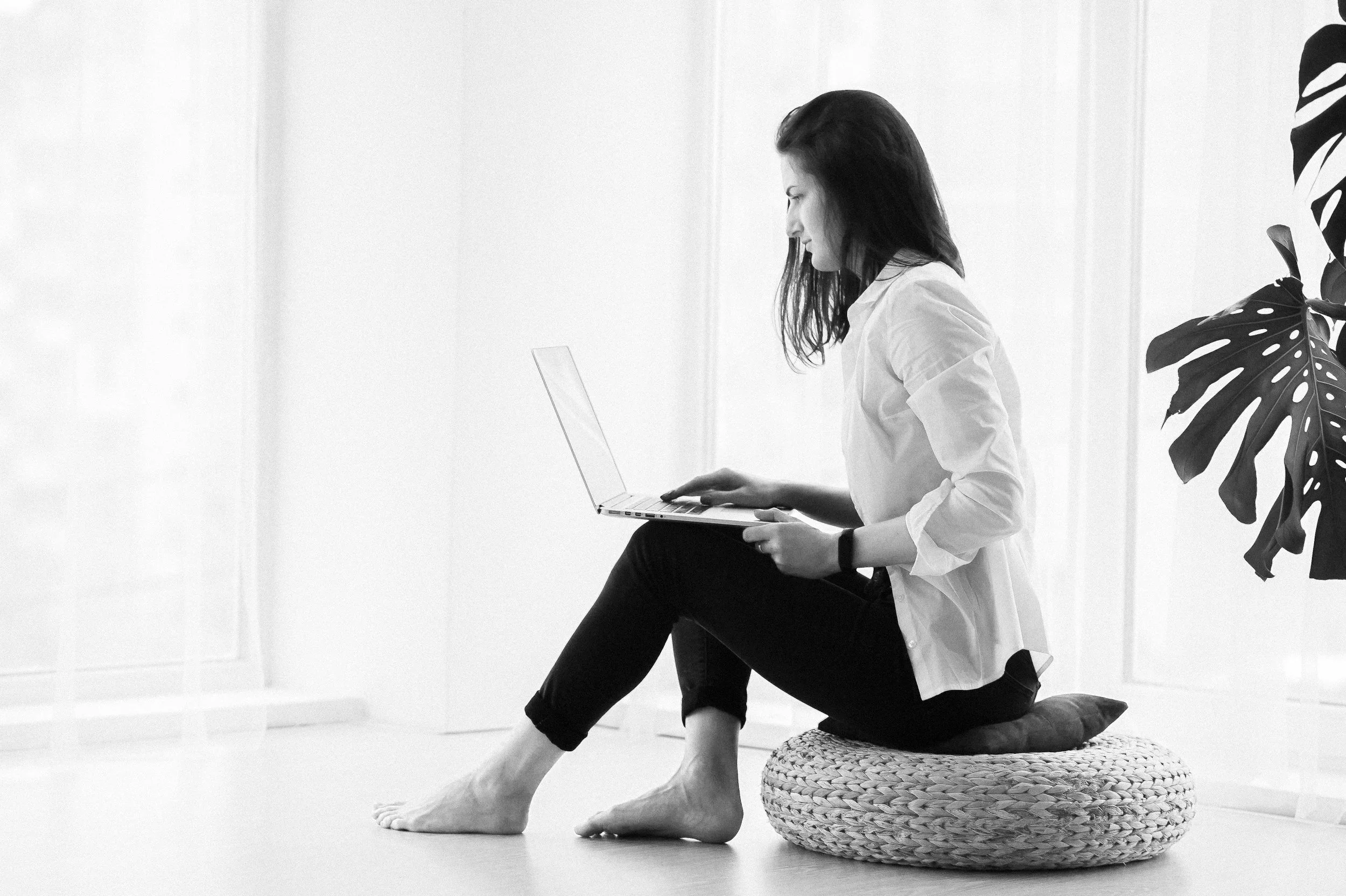 A woman sitting on a basket, using a laptop, kneeling on a cushion, with a large plant behind her, in a room with blank walls.