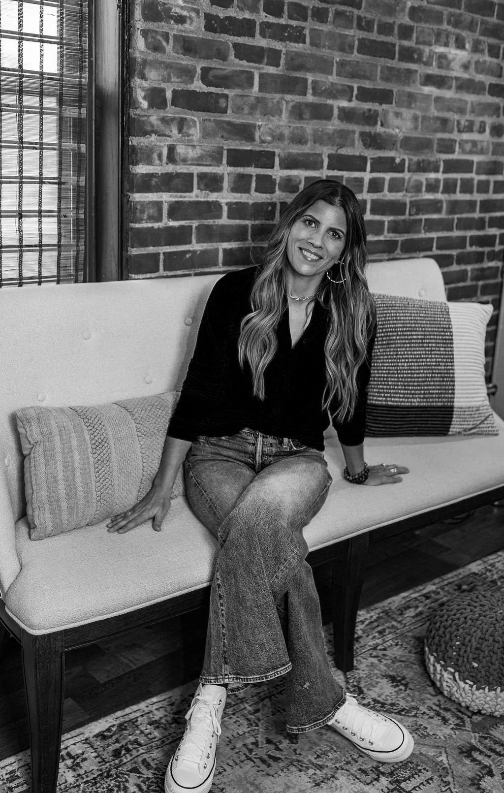A woman with long wavy hair sitting on a sofa in front of a brick wall, smiling at the camera. She is wearing a black top, jeans, and white sneakers, accessorized with hoop earrings and a necklace.