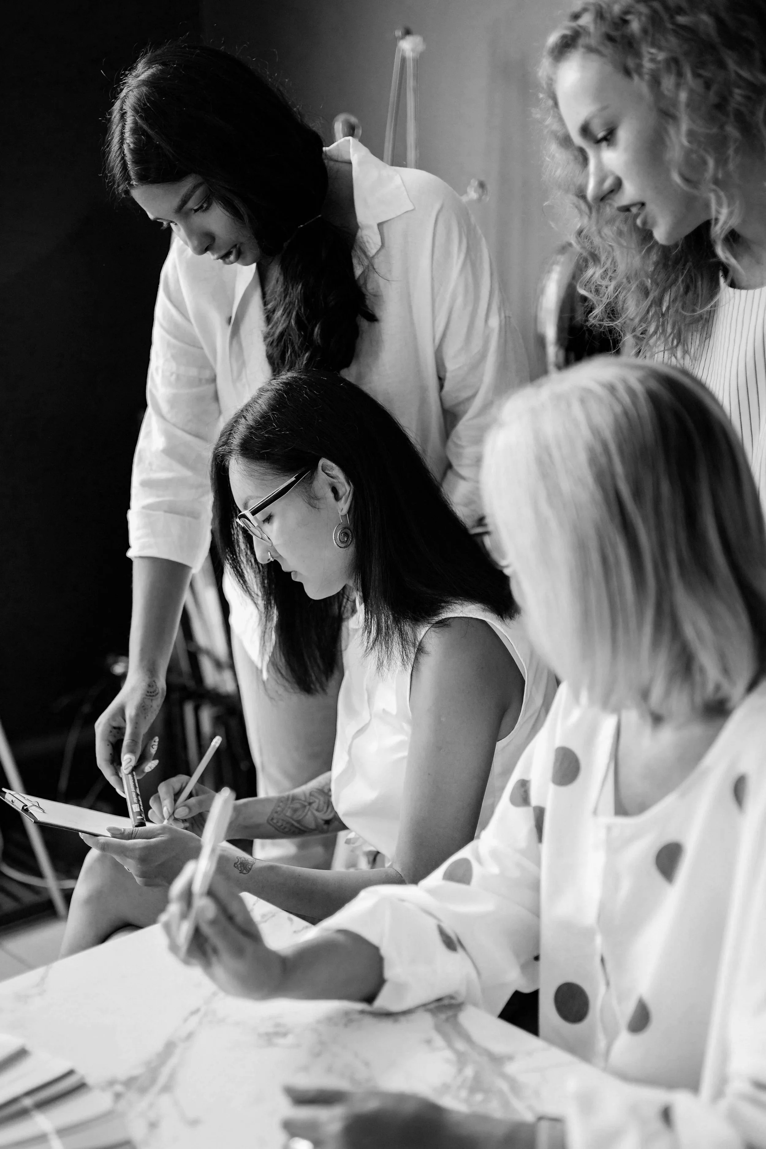 Two women are focused on writing or drawing on a clipboard at a table, with a third woman sitting nearby, all engaged in a creative or collaborative activity.