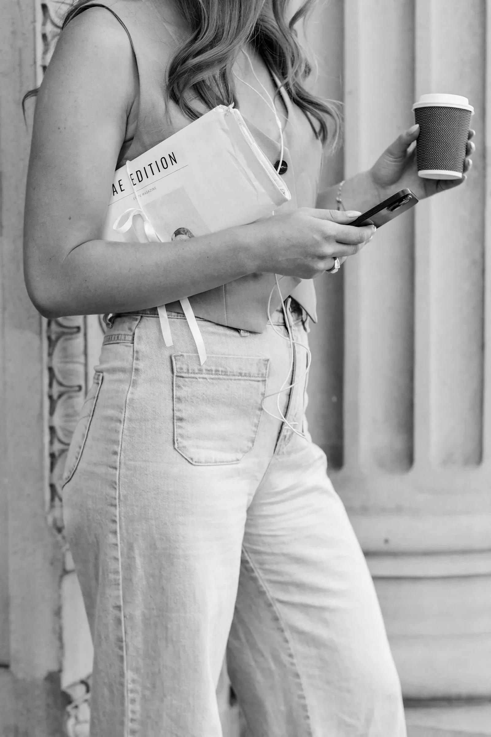 A woman holds a coffee cup and a smartphone, with a magazine tucked under her arm. She is wearing a sleeveless top and jeans and is listening to earphones.