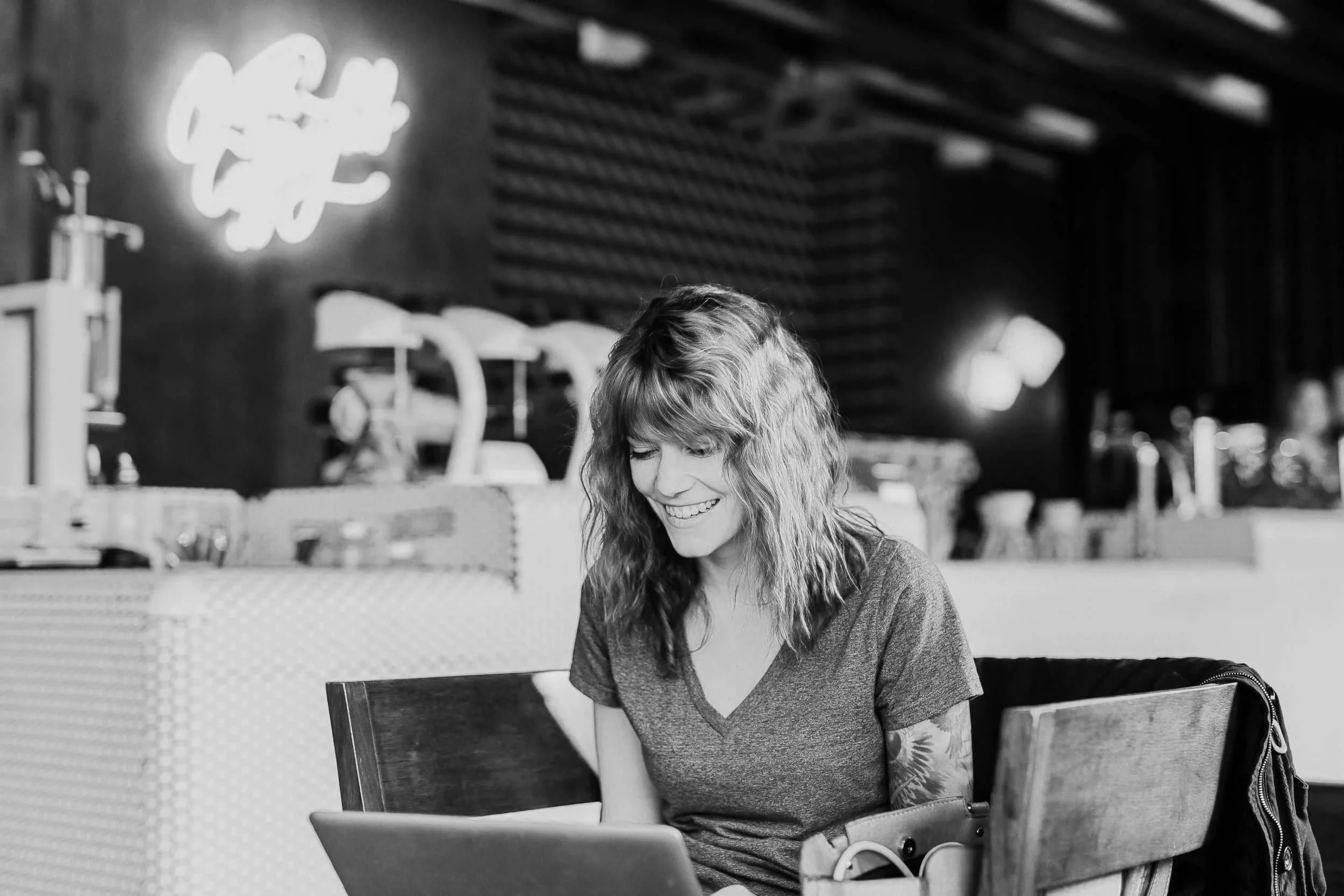 ADHD coaching for Women virtual with wavy hair smiling while accessing virtual ADHD support on her laptop in a modern café with a neon sign in the background.