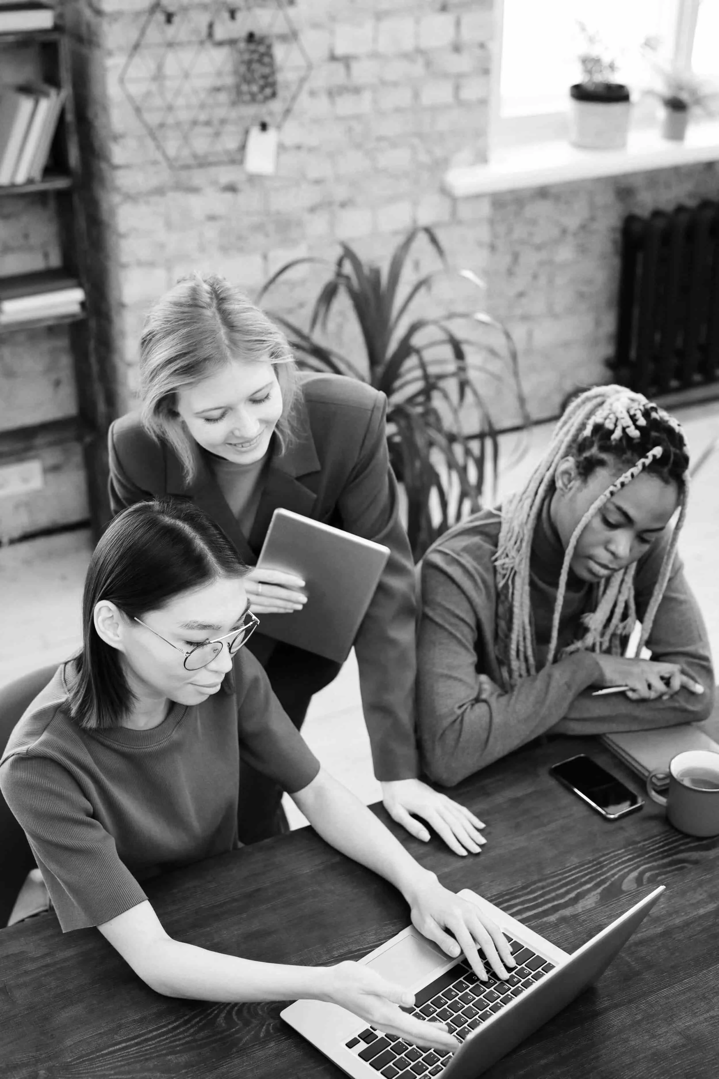 Three women collaborate at a desk with a laptop, notebook, and phone in a modern office setting.