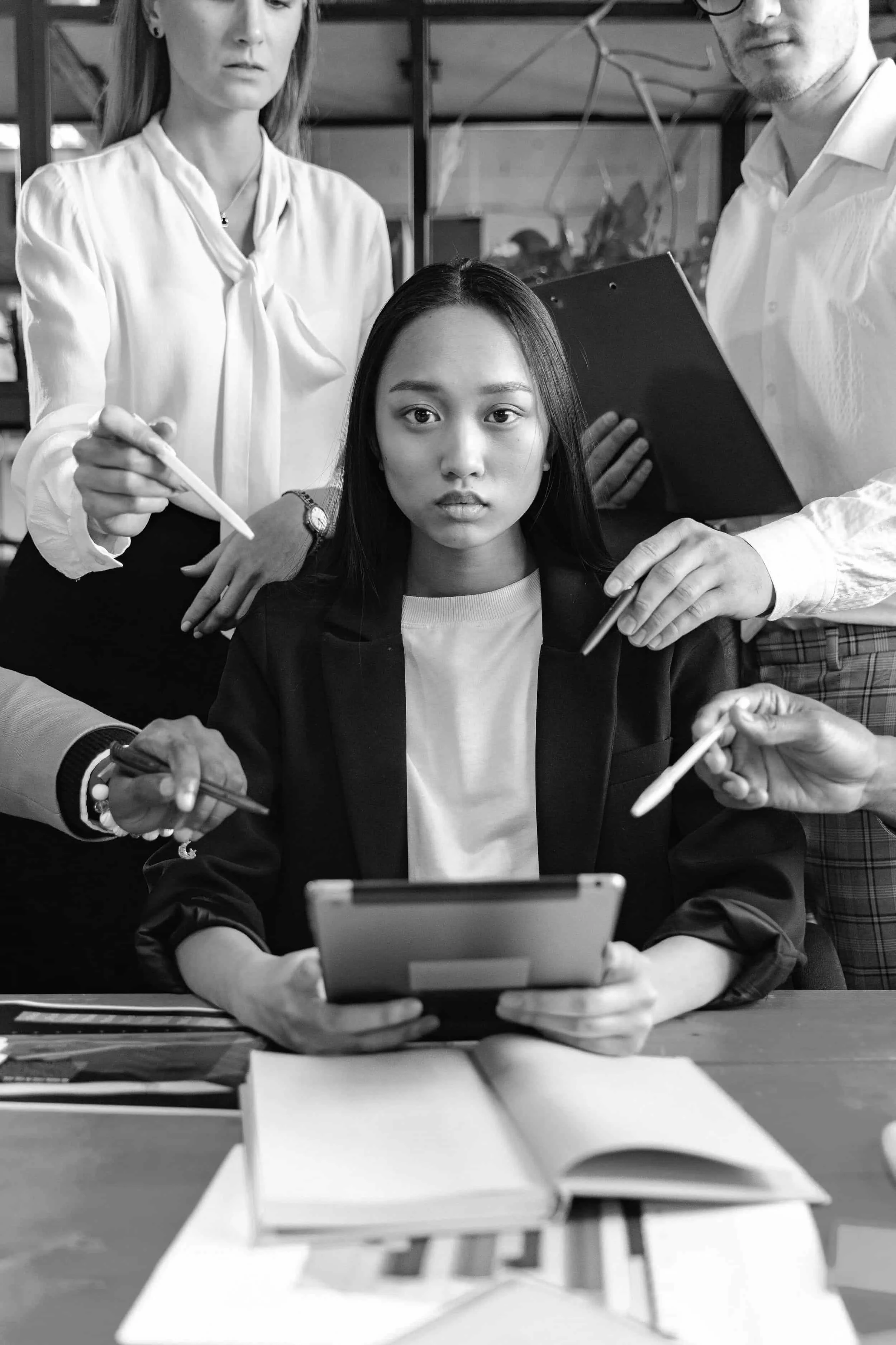 A woman sits with a tablet, surrounded by people pointing pens at her, looking overwhelmed.