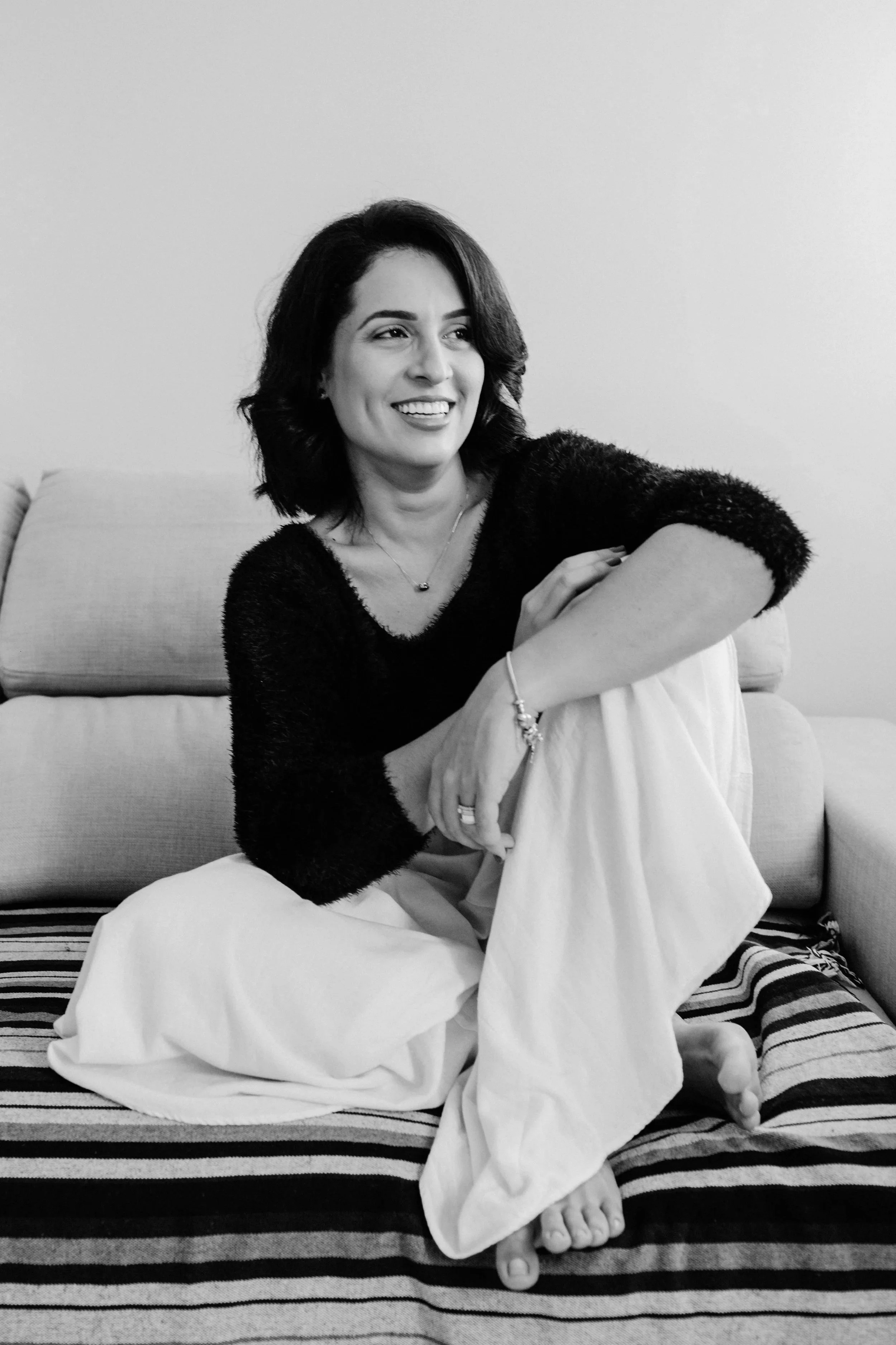 A woman with short dark hair wearing a black sweater and white pants sitting on a bed with a striped blanket, smiling and looking to the side in a black-and-white photo.