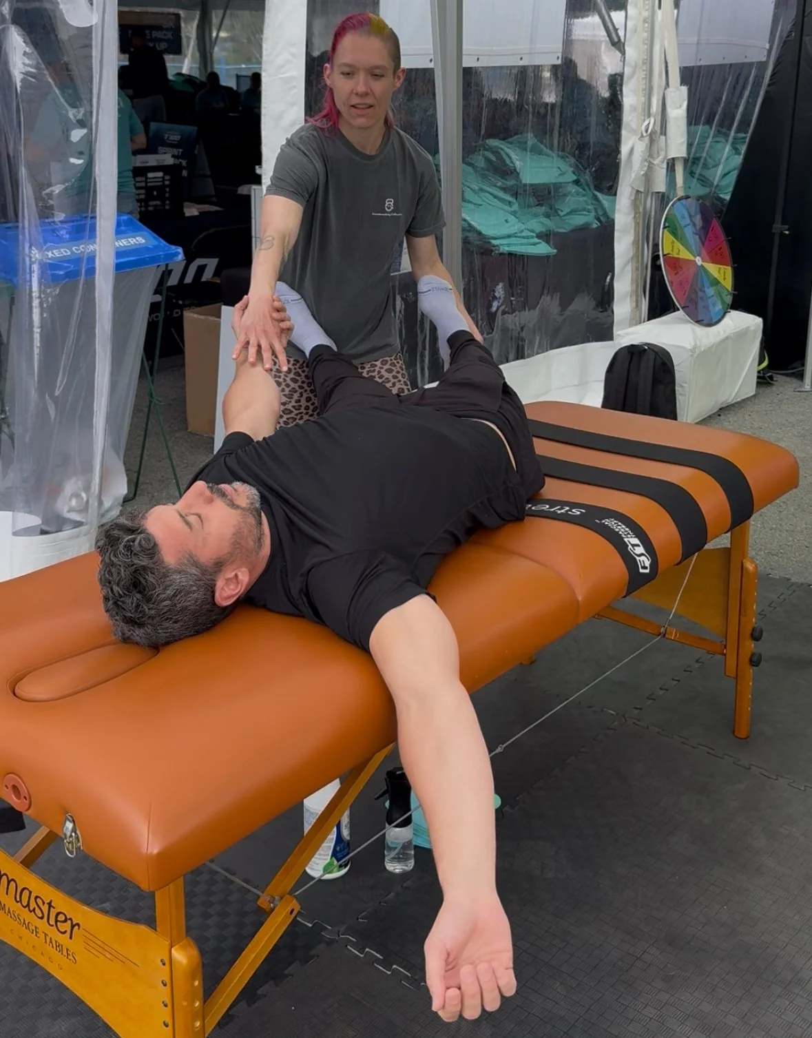 A woman providing a massage to a man lying face-up on an orange massage table outdoors under a tent. The woman has pink hair and is wearing a gray shirt, and the man has gray curly hair and is wearing a black shirt.
