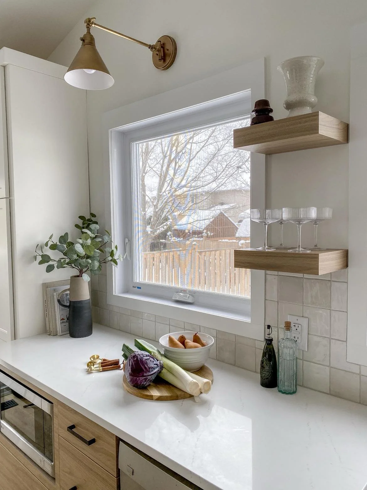 Kitchen countertop with a bowl of pears, purple cabbage, leek, and green onion; window with outside view of a snowy yard and trees; decorative vases and glassware on wooden shelves; potted plant on counter.