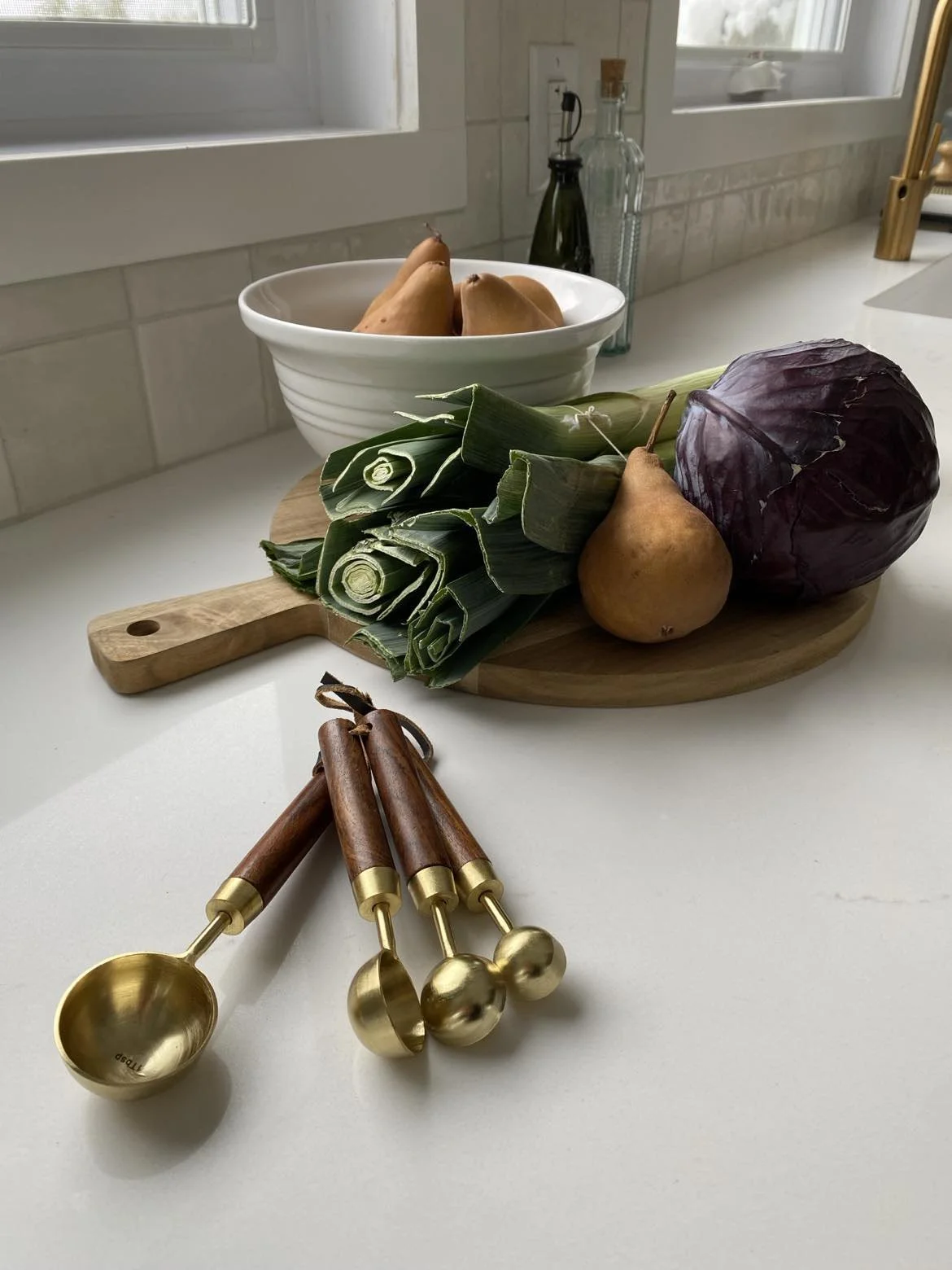 A kitchen countertop with a cutting board holding leeks, a pear, and a head of red cabbage, a white bowl with pears, and four gold measuring spoons with wooden handles.