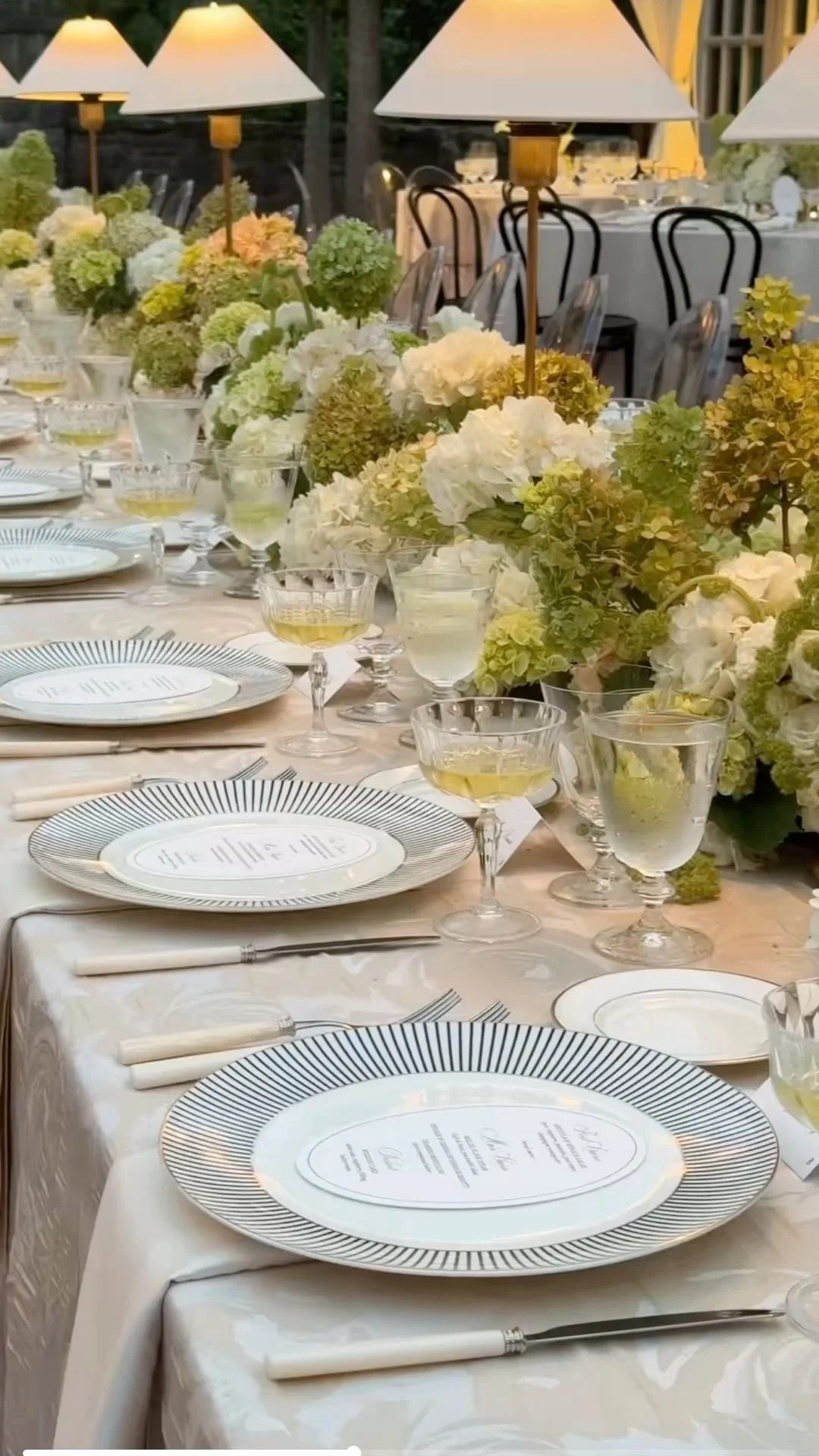 A beautifully set outdoor banquet table with white floral centerpieces, gold lamps, and elegant place settings including white plates with black lines, yellow-colored drinks, and glassware.