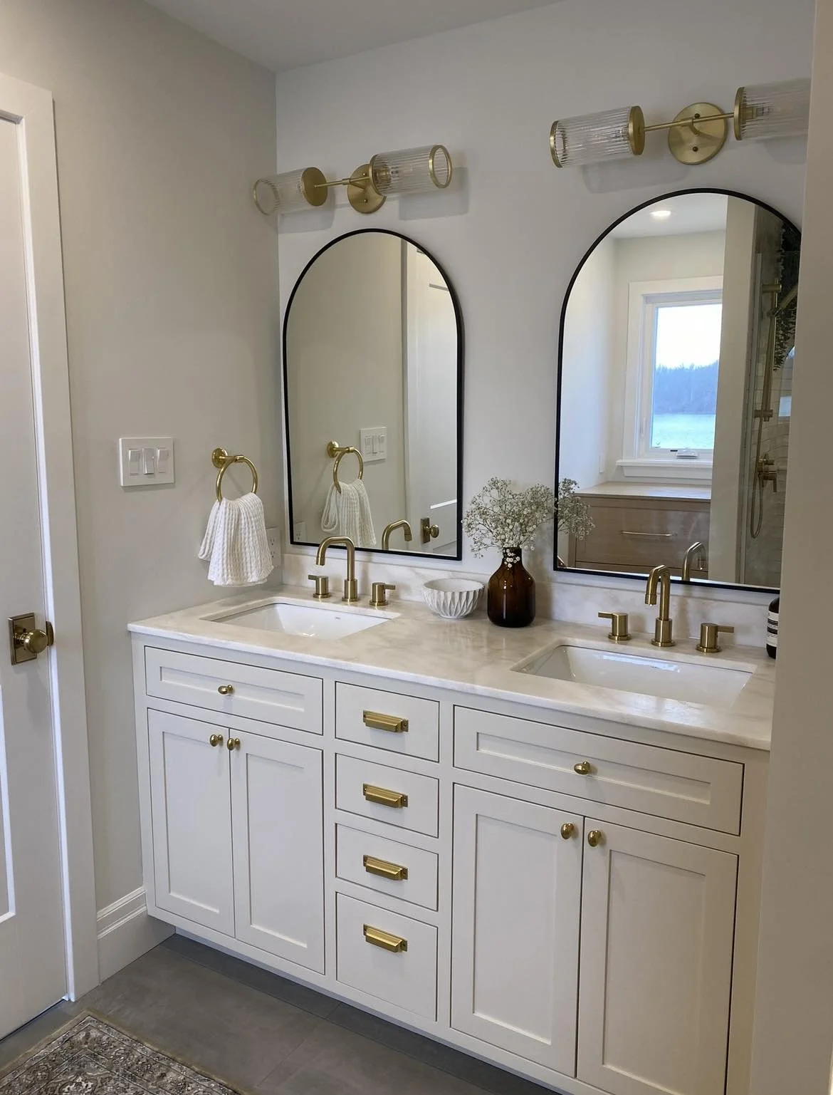 A modern bathroom vanity with white cabinets, gold hardware, two sinks, two arched mirrors, and two wall-mounted light fixtures with glass shades. Decor includes a brown vase with dried flowers and a white bowl.