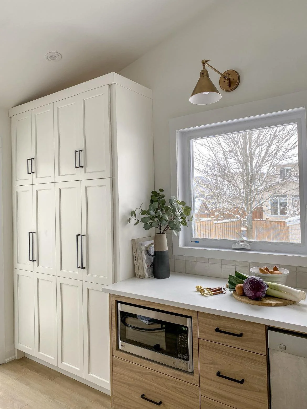 A kitchen with white cabinets, a window showing a snowy outdoor scene, and a countertop with vegetables and a potted plant.