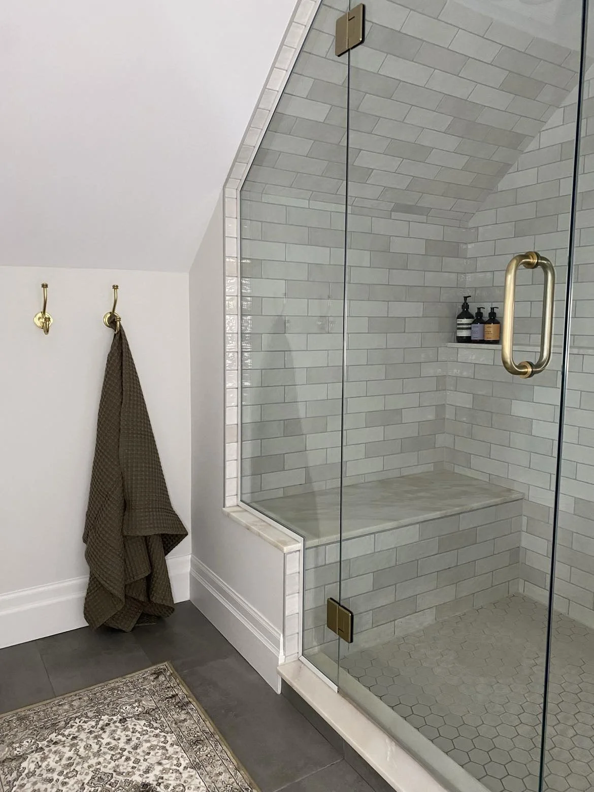 A modern bathroom featuring a glass-enclosed shower with white subway tiles, a brass handle, and a built-in bench. There are two black bottles on a shelf inside the shower, and two gold hooks on a white wall outside, with a brown towel hanging on one hook. Part of a patterned rug and dark gray tile flooring are also visible.