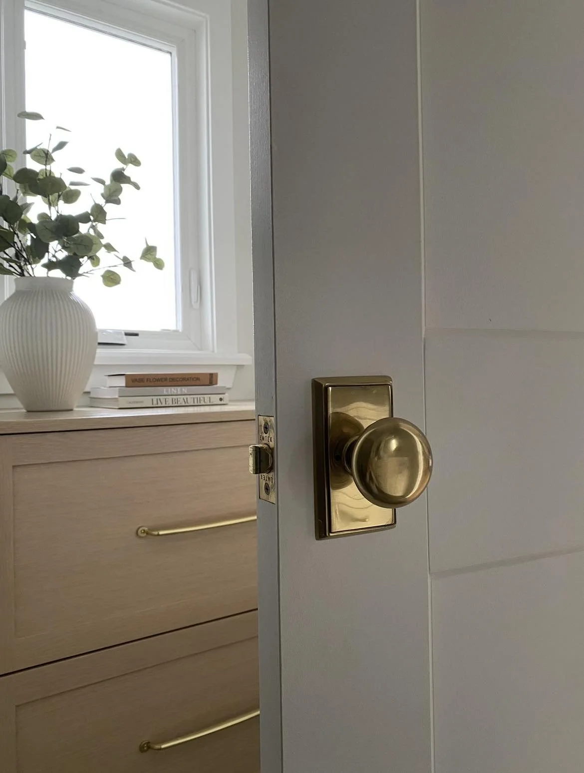 Partially open white room door with brass doorknob and lock, leading to a bedroom with a light wood dresser, white window, white vase with green leaves, and stacked books on the windowsill.
