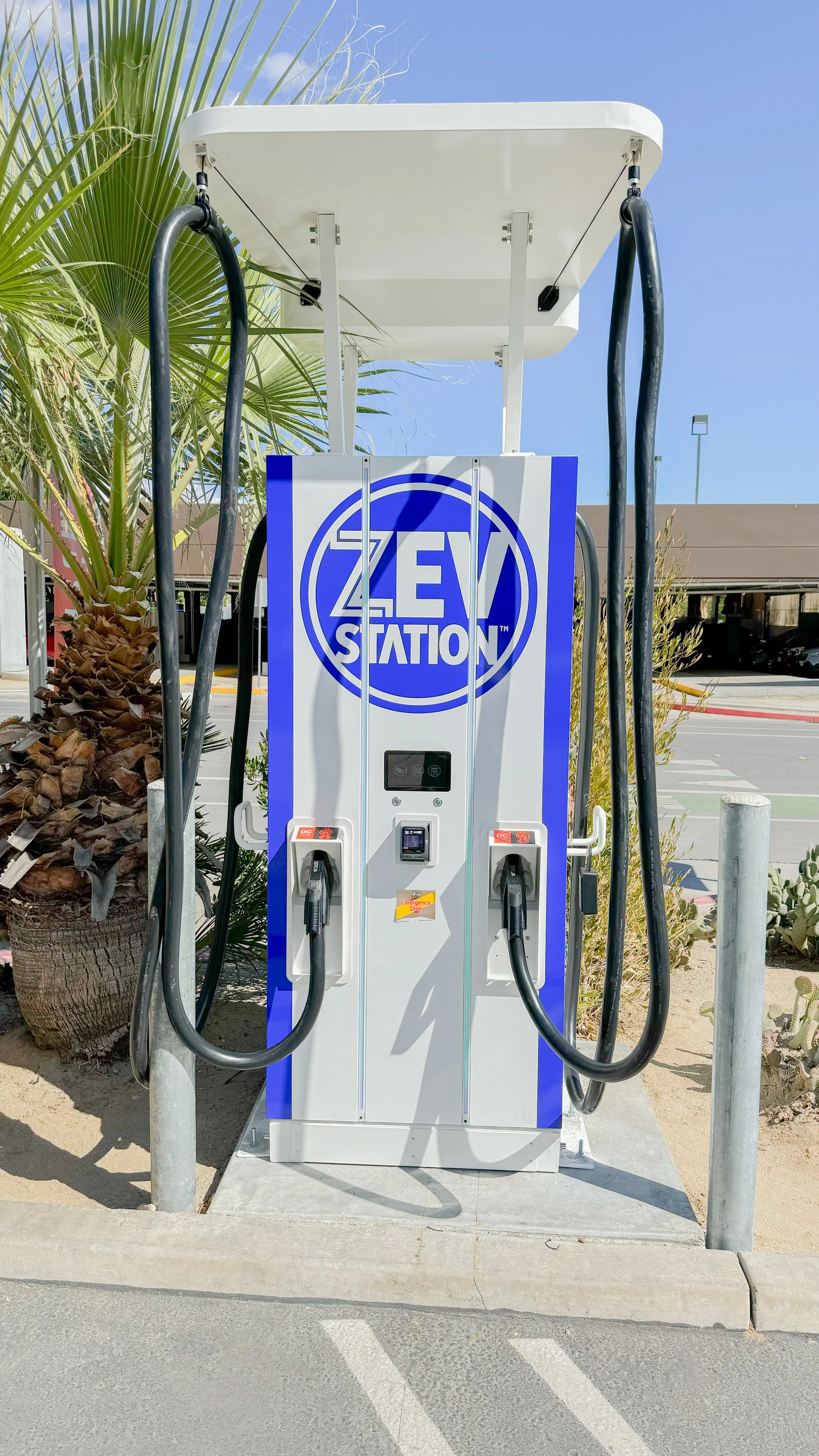 Electric vehicle charging station with a white and blue design, two charging cables, and a digital display, located outdoors next to desert plants and a parking lot.