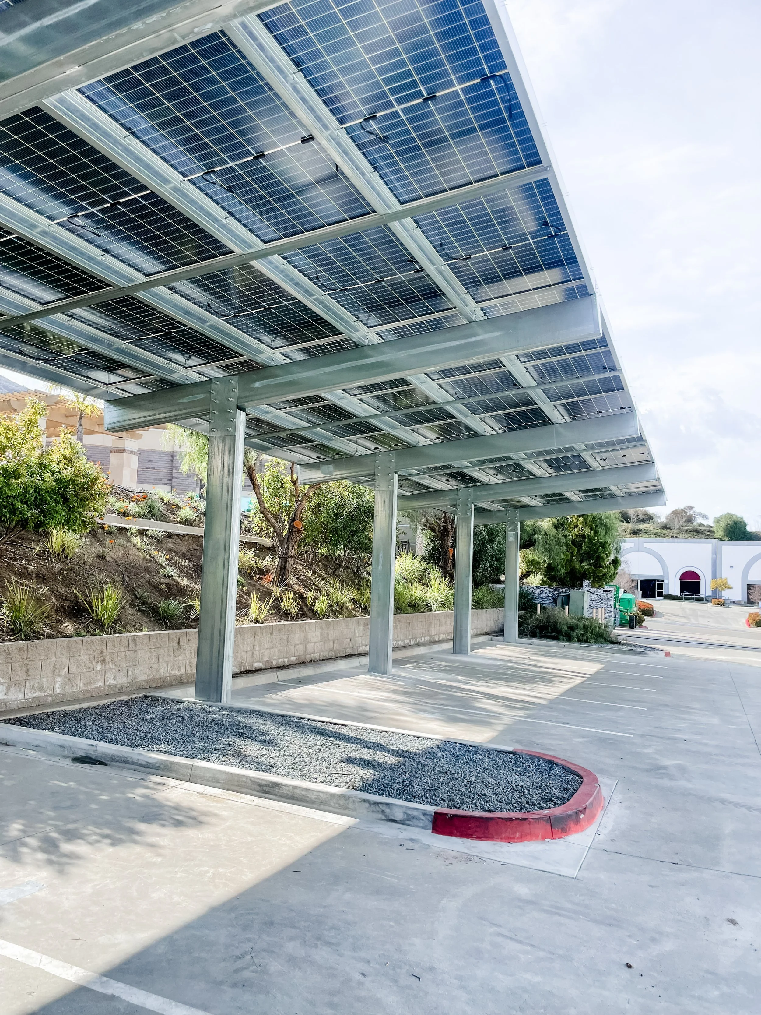 Solar panels installed on a metal canopy over a parking lot, with a landscaping area and a building in the background.