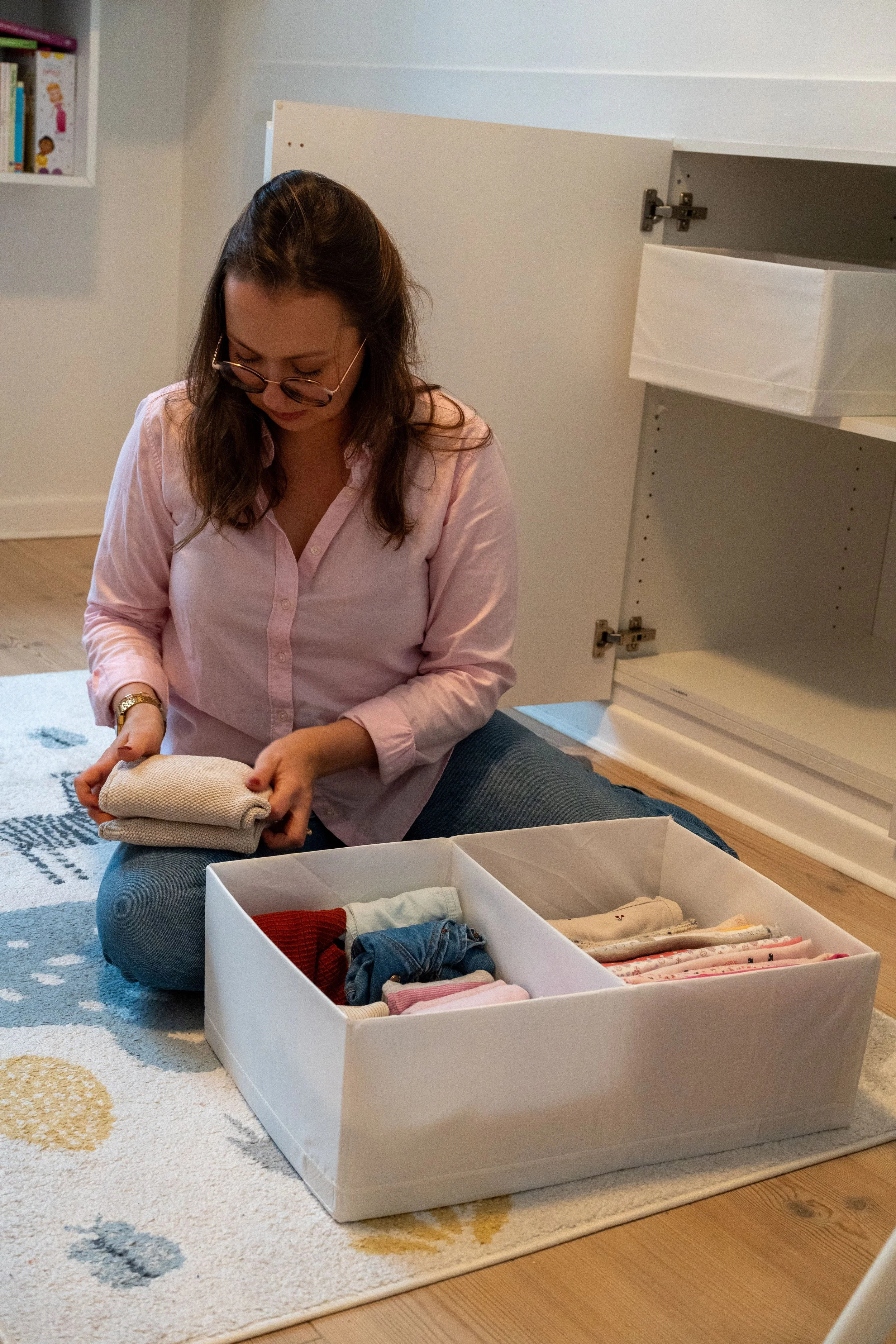 A woman with glasses and a pink button-down shirt is sitting on the floor, folding clothes. In front of her, there is a white storage box filled with neatly folded clothes and folded laundry on top. She is in a room with a patterned rug and open white cabinets behind her.