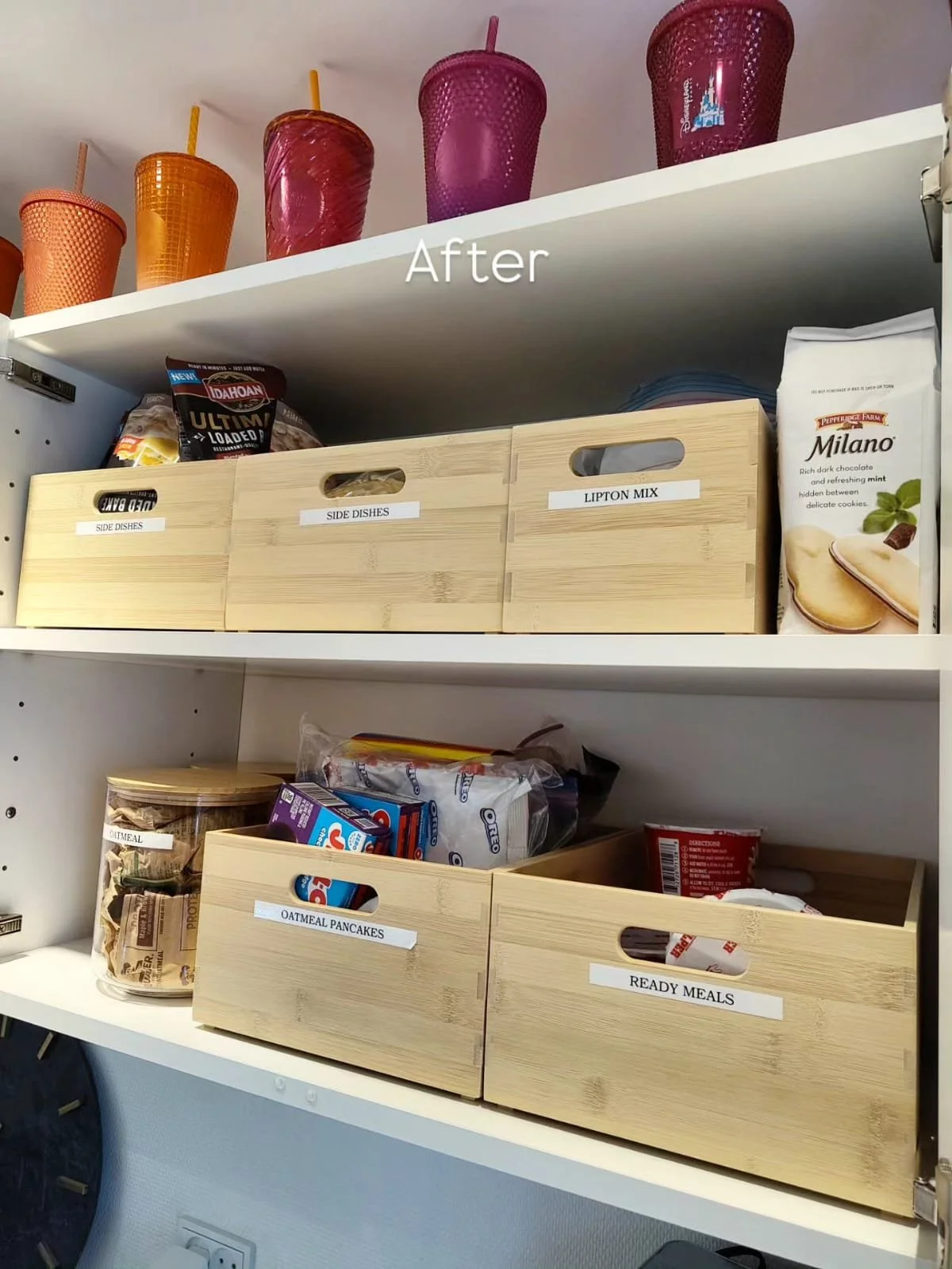 White kitchen shelf with four pastel-colored cups on the top, labeled storage boxes on the middle and lower shelves, including one labeled 'Oatmeal Pancakes' and another 'Ready Meals', and a bag of snack chips.