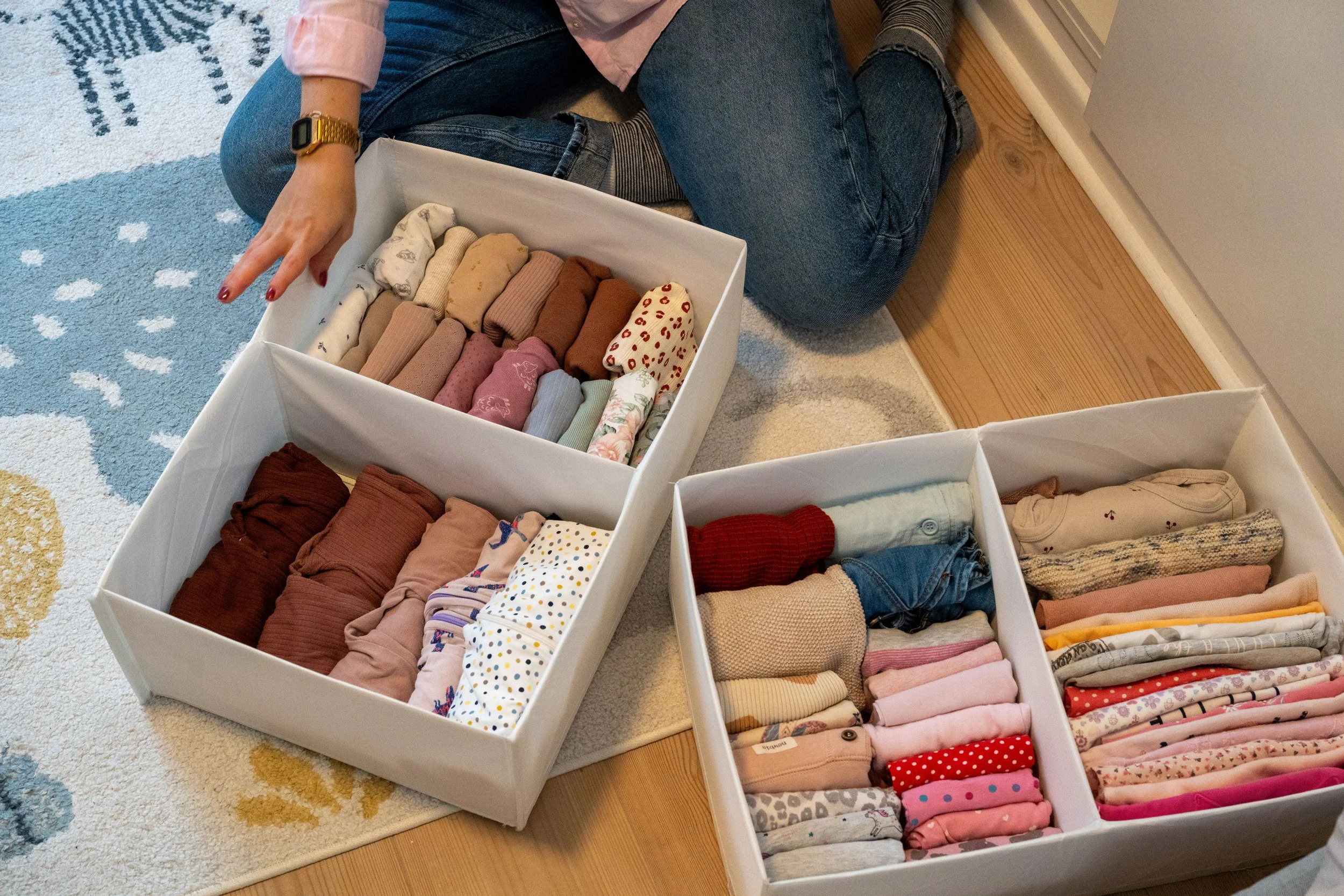 A person sitting on the floor with three white storage boxes filled with folded clothes, including socks, shirts, and other garments, nearby a colorful carpet and wooden floor.