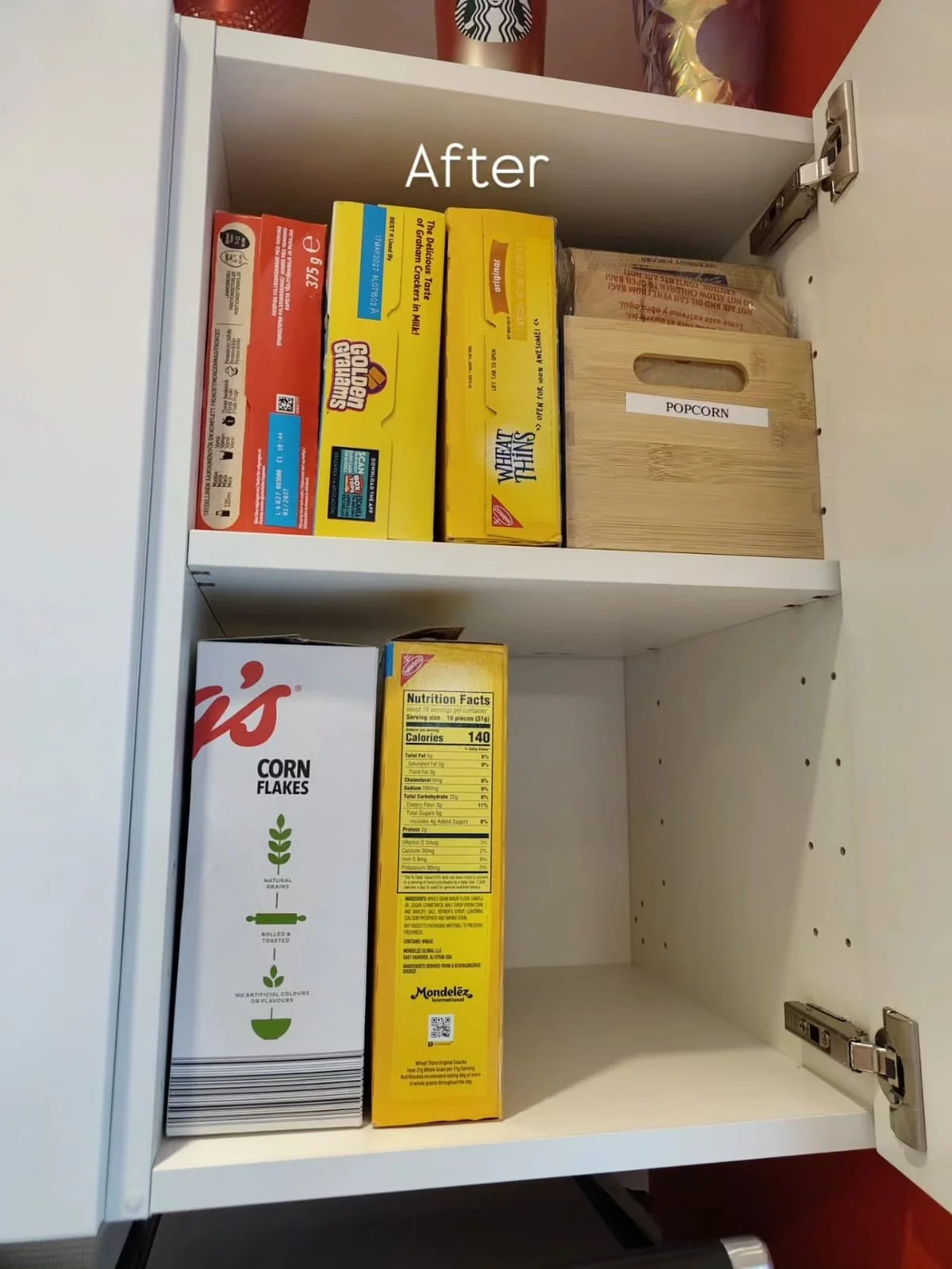 A white kitchen cabinet with two shelves, containing cereal boxes, a wooden popcorn box, and a box of cornflakes.