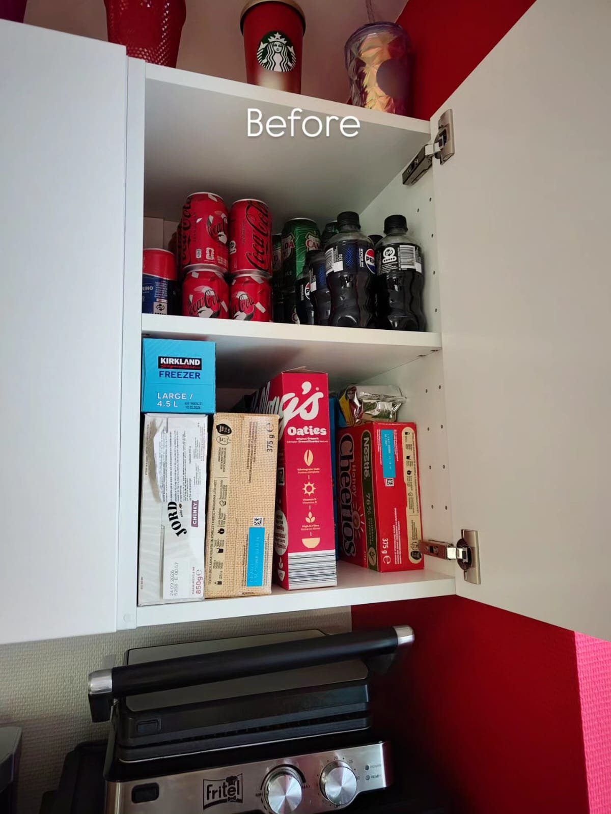 Open kitchen cabinet with soda cans, bottles of soda, and snack boxes inside. The cabinet door is white, and there is a red wall in the background.