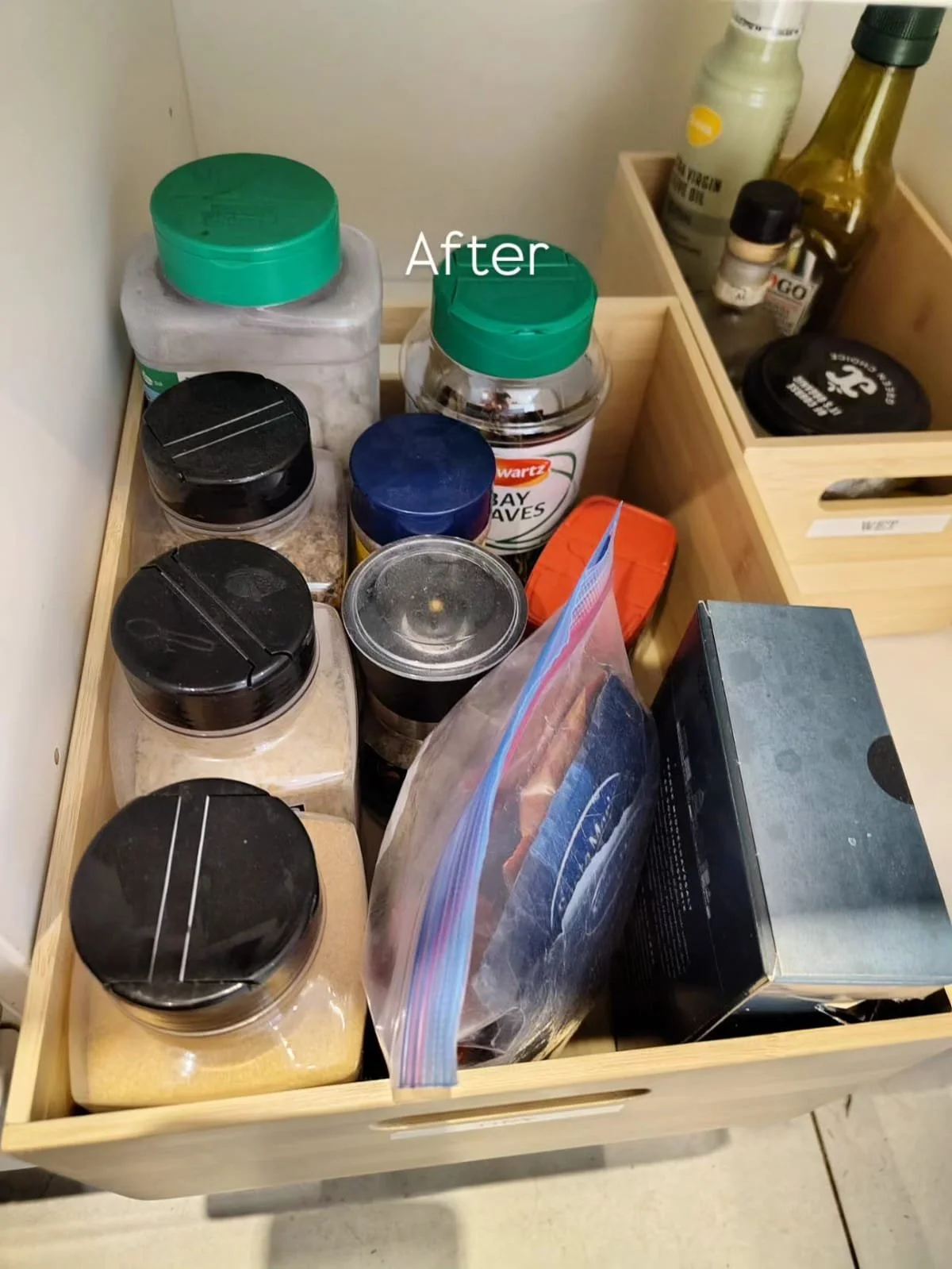 A wooden drawer filled with various spices, seasonings, and containers, with some in jars and others in plastic bags, and a small section of another container with condiments visible.