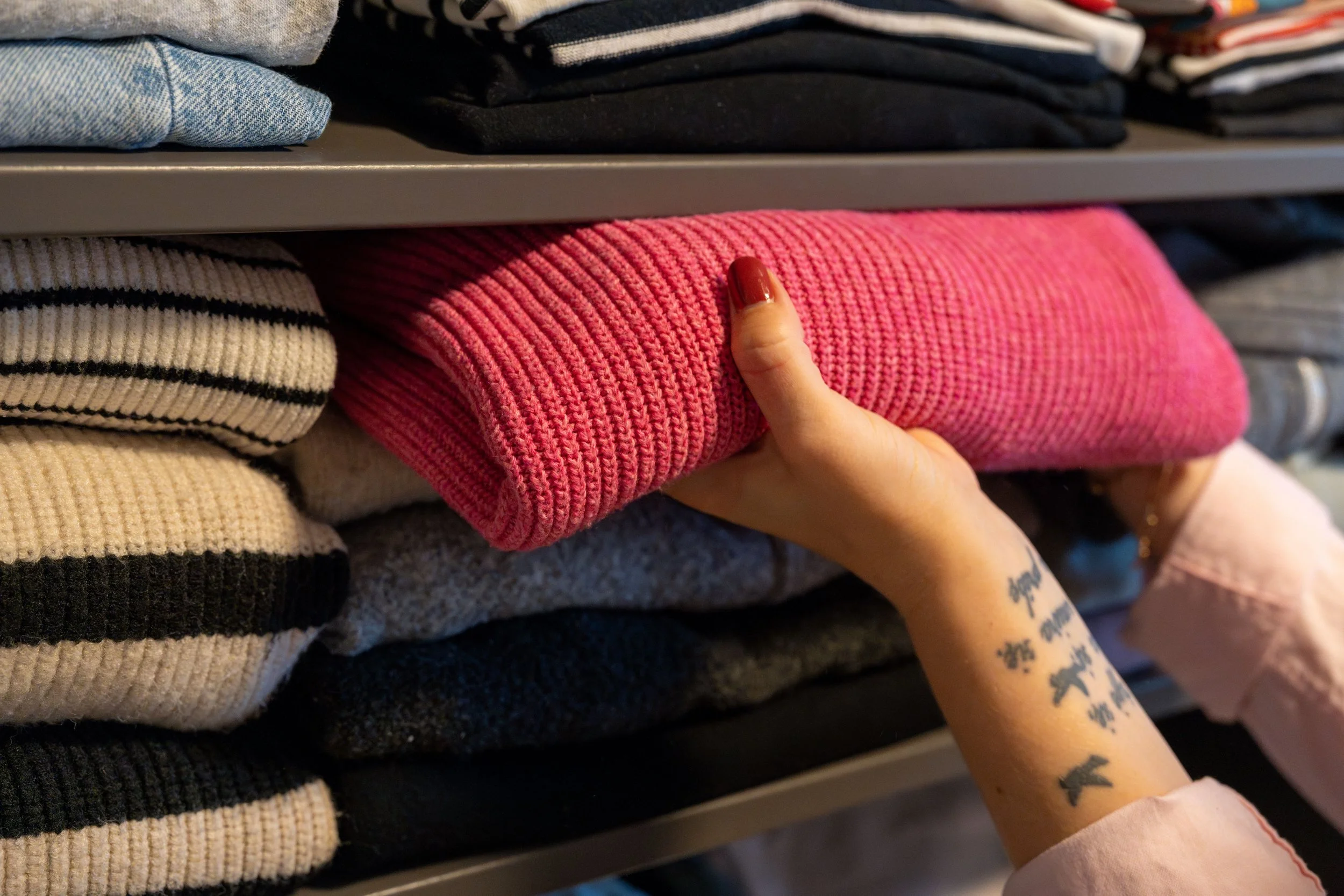 A hand with tattoos pulling out a bright pink knit sweater from a pile of folded clothes on a shelf. The shelves contain various folded garments, including striped, denim, and dark-colored pieces.