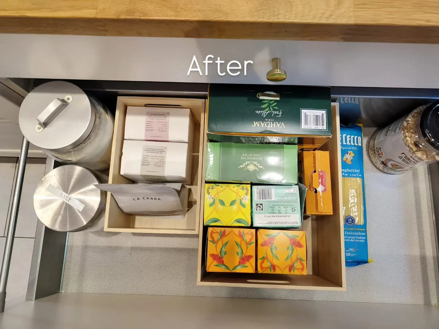 Open kitchen drawer containing salt and sugar containers, tea and pasta boxes, and a jar of grains.