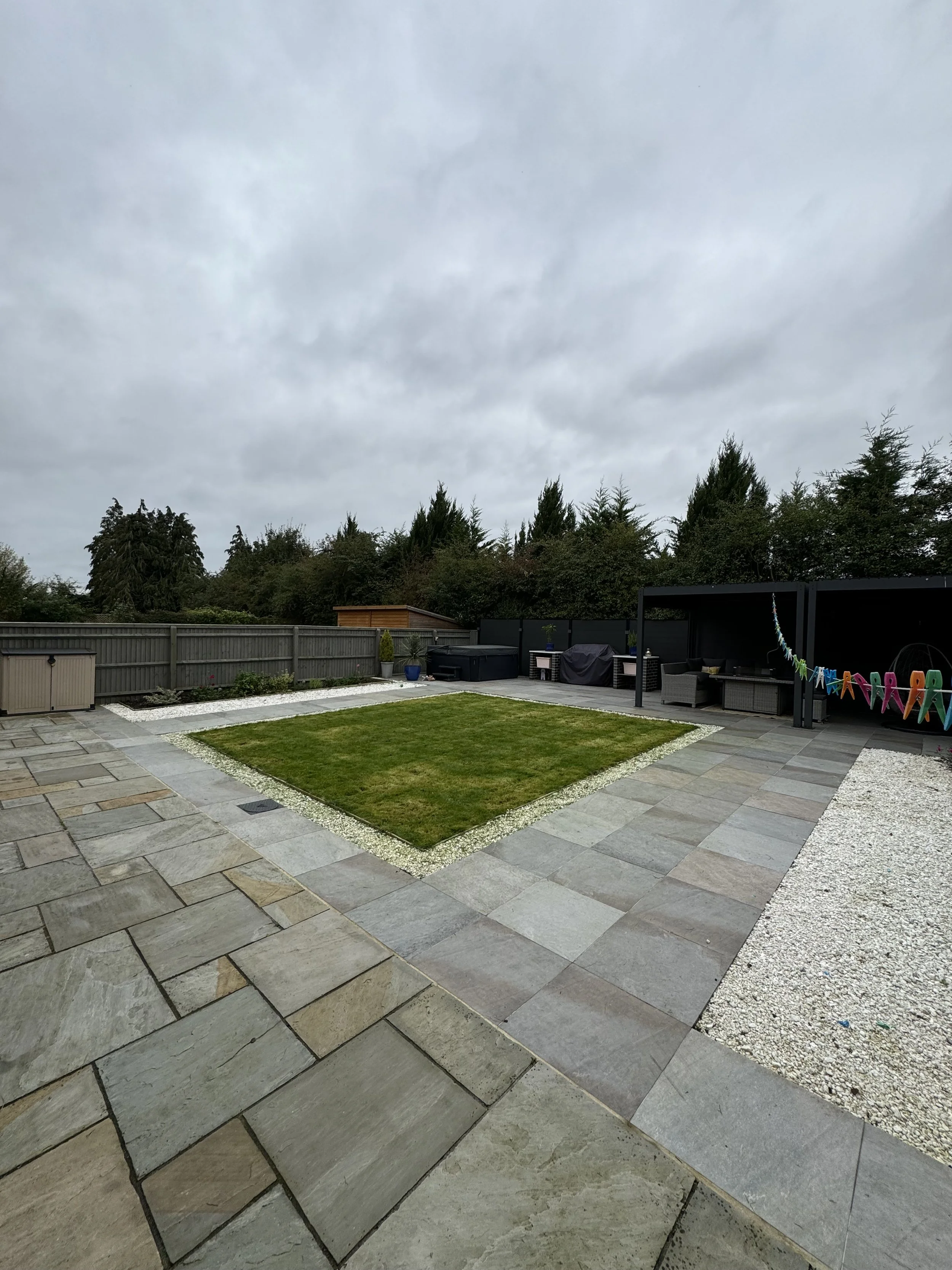Backyard with stone patio, small grassy area, gravel borders, trees in background, and outdoor furniture under covered area.