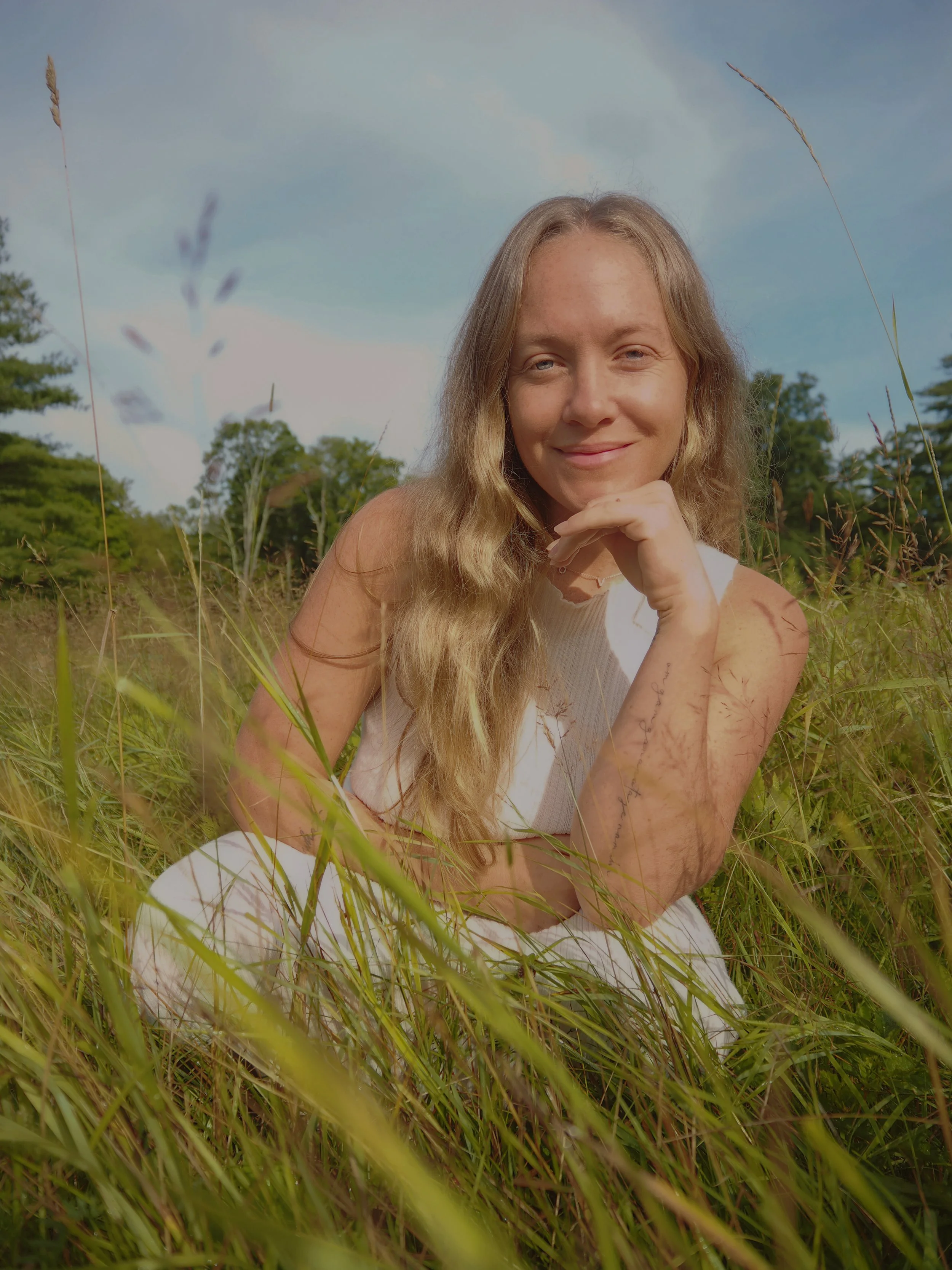 A woman with long blonde hair sitting in a grassy field, smiling and resting her chin on her hand, with a blue sky and trees in the background. Dru Erin operations and clarity strategist for wellness businesses